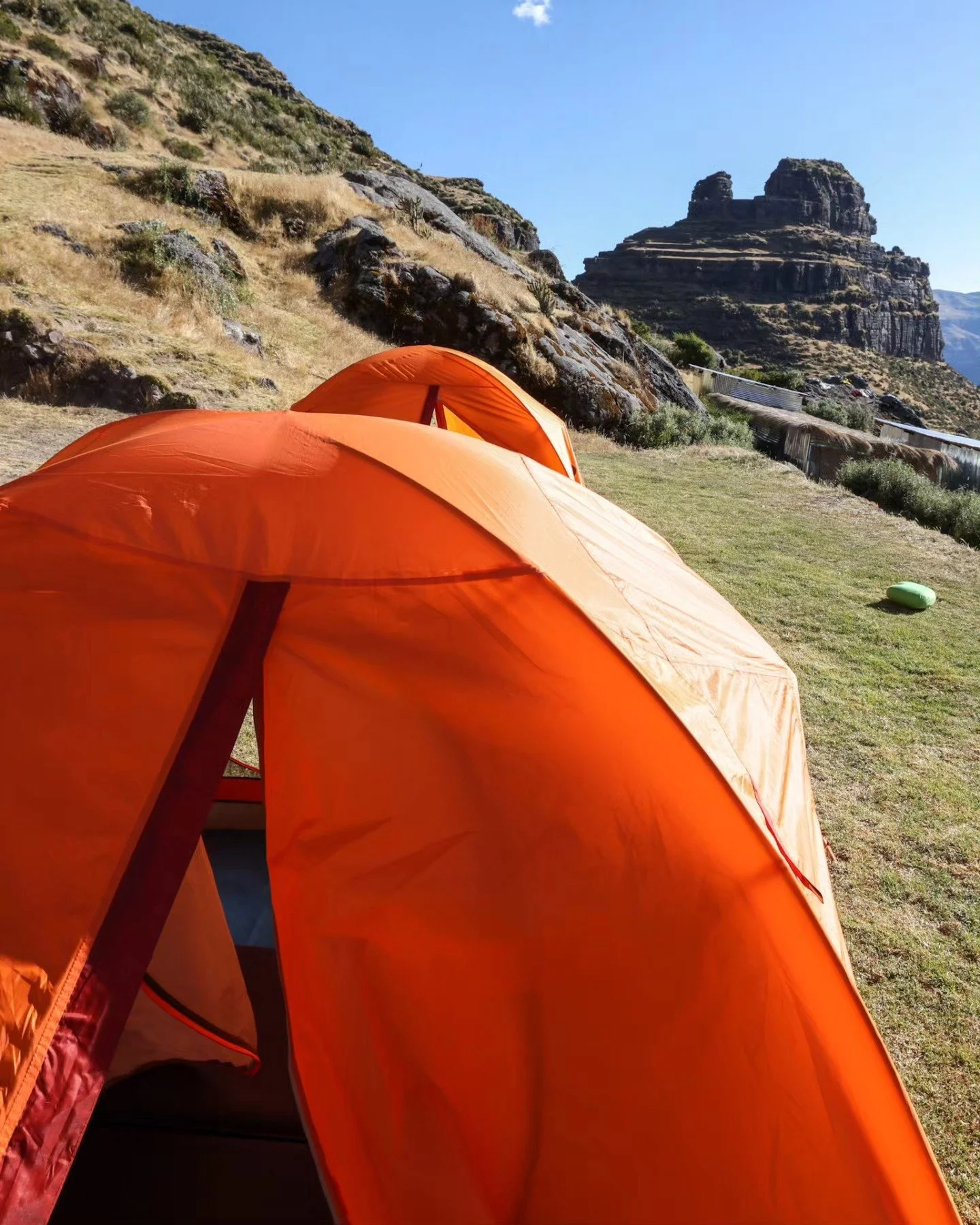 Two orange camping tents set up on a grassy area with rocky hills and a unique layered rock formation in the background under a clear blue sky.