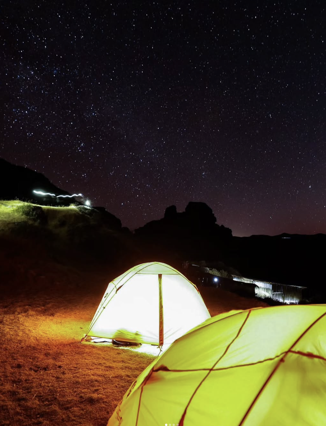 Nighttime scene of a camping site with glowing yellow tents on sandy ground under a star-filled sky, with hills or mountains in the background.