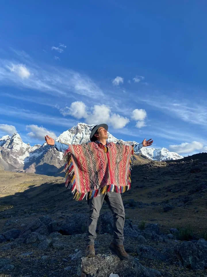 A man standing on rocks in a mountainous landscape with snow-capped peaks in the background, wearing a wide-brimmed hat and a colorful patterned poncho, arms outstretched, facing the sky.