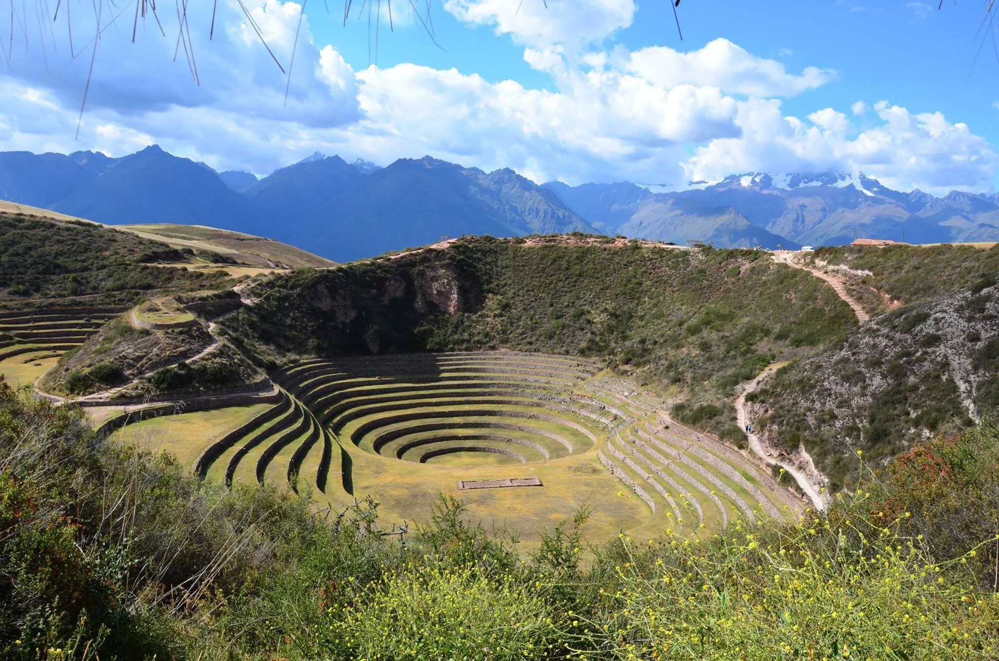 Inca archaeological site of Moray with circular terraced agricultural terraces, surrounded by green hills and mountains under a partly cloudy sky.