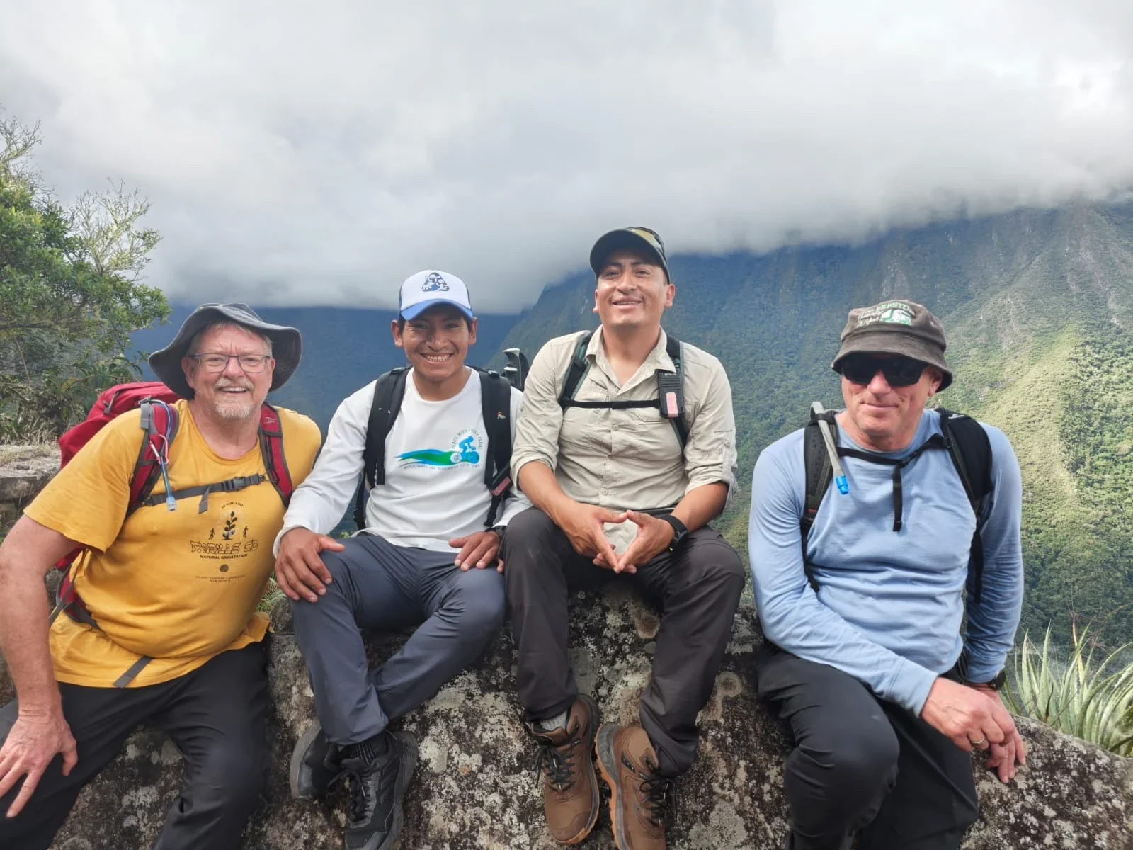 Four men sitting on a rock in a mountainous area with lush green vegetation and cloudy sky in the background, dressed in hiking clothes and backpacks, smiling at the camera.