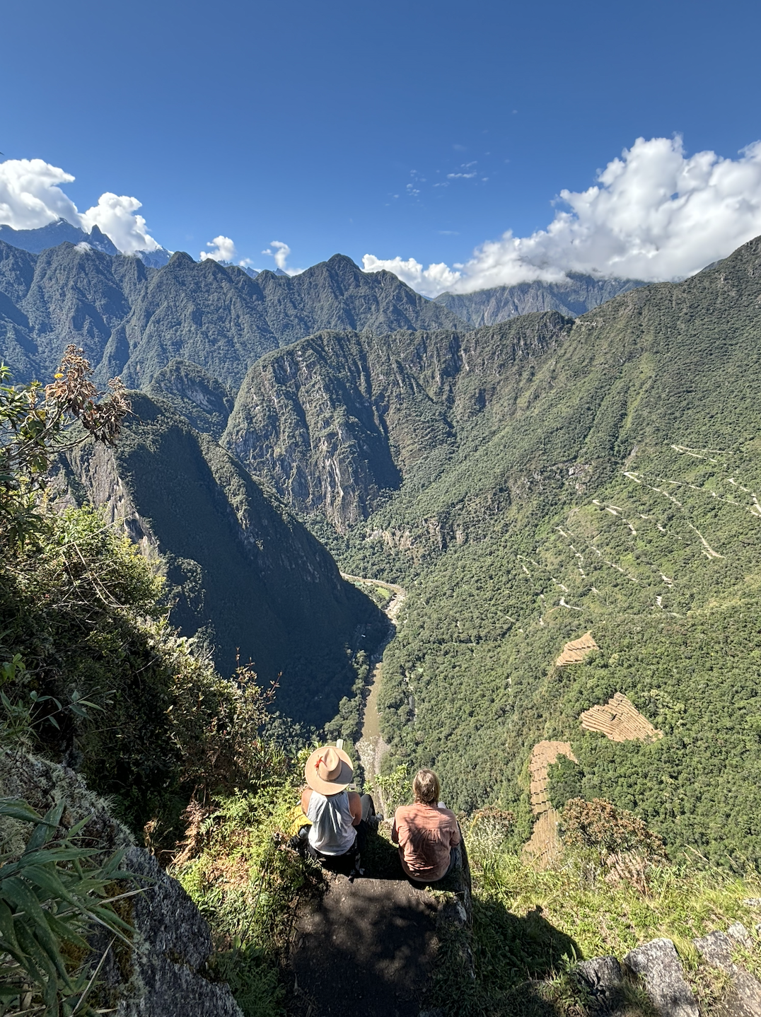 Two people sit on a rocky ledge overlooking a deep green valley with tall mountains, lush forests, and a river below, under a blue sky with some clouds.