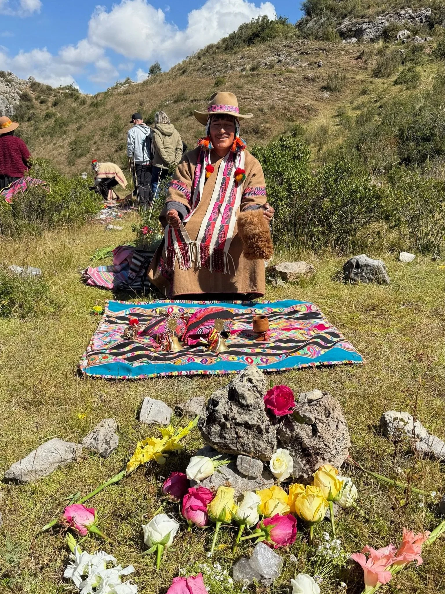 A woman dressed in traditional Andean clothing stands outdoors in a grassy area with rocks and shrubs, holding a furry object. In front of her is a colorful cloth with small decorative items, and an arrangement of multicolored roses and rocks.