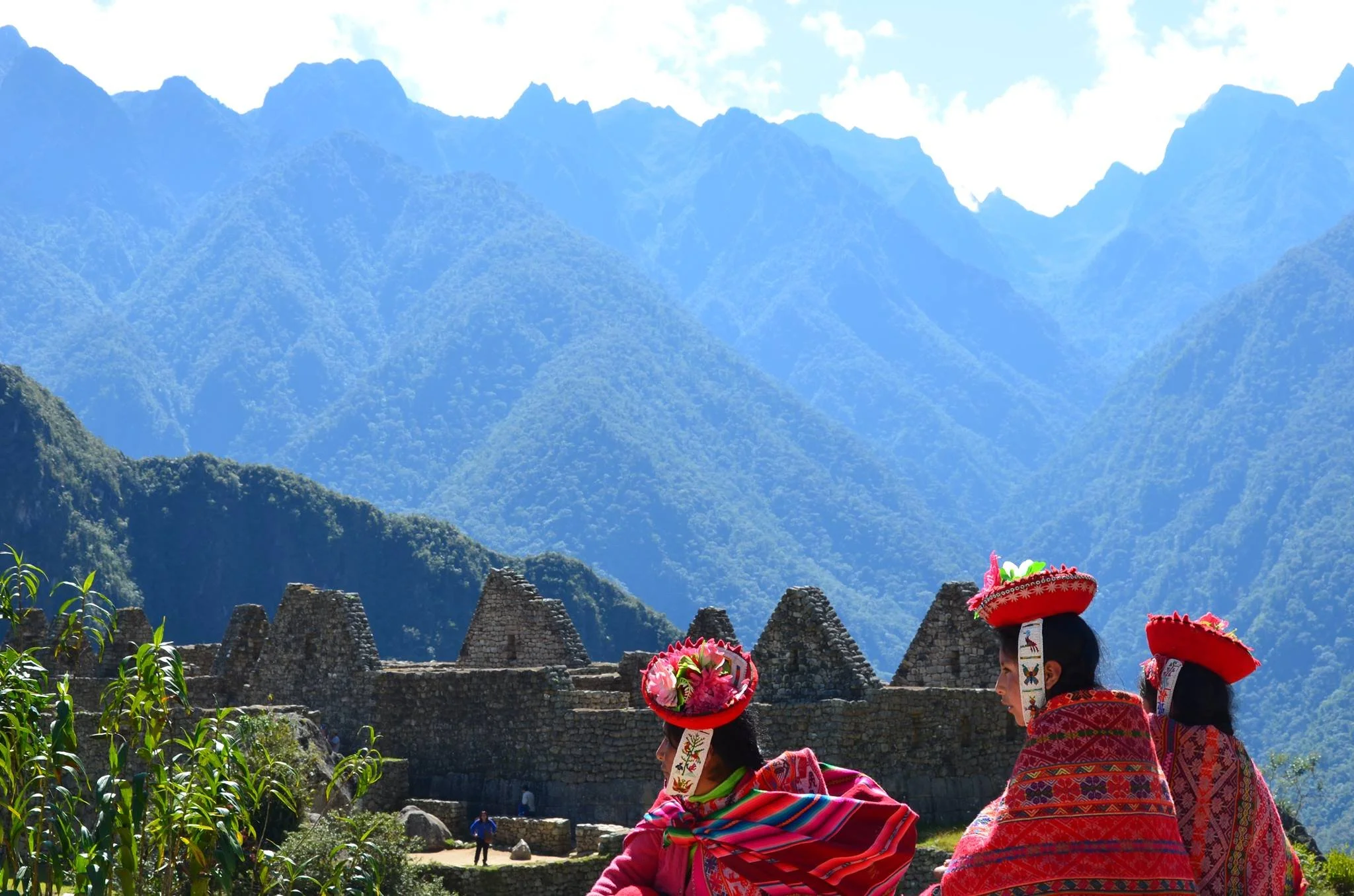 Three indigenous women dressed in traditional colorful clothing and hats, sitting in front of an ancient stone structure with a mountainous landscape in the background.
