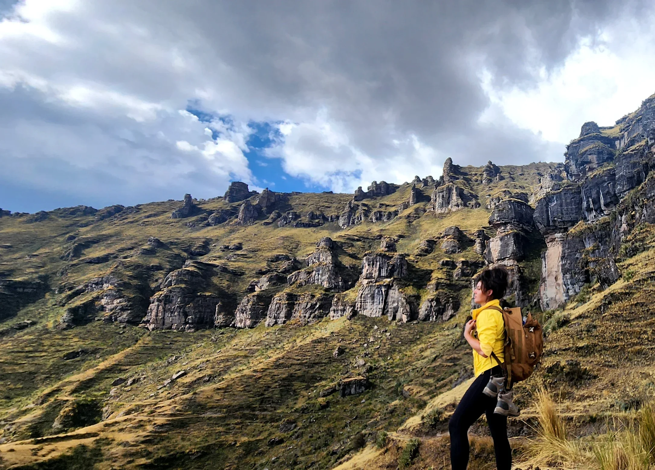 A woman hiking in a mountainous area with rocky cliffs and grass, wearing a yellow jacket and carrying a backpack, under a cloudy sky.
