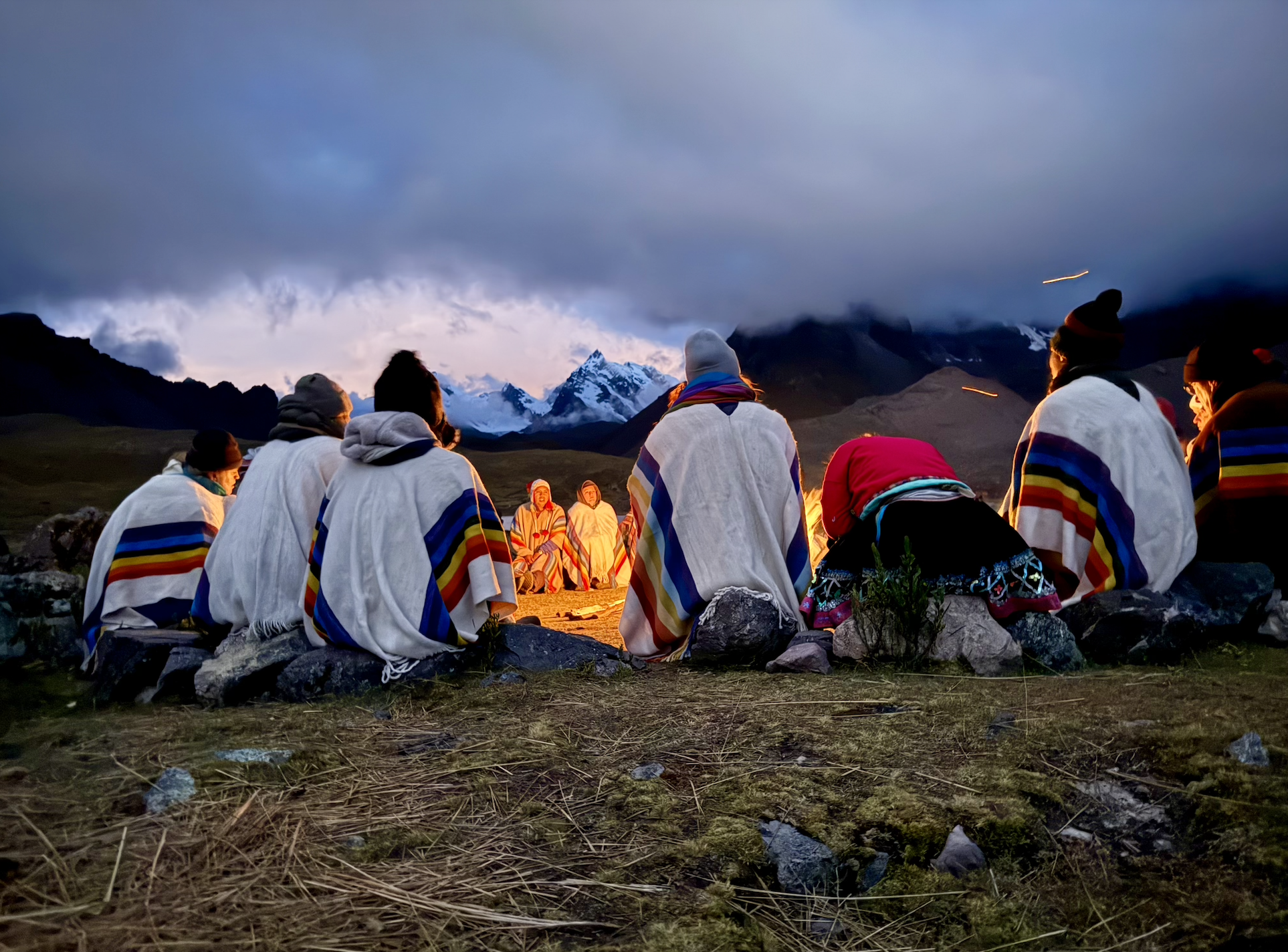 A group of people sitting around a campfire on a mountain plain with snow-capped peaks and dark clouds in the background, wearing traditional Andean clothing.