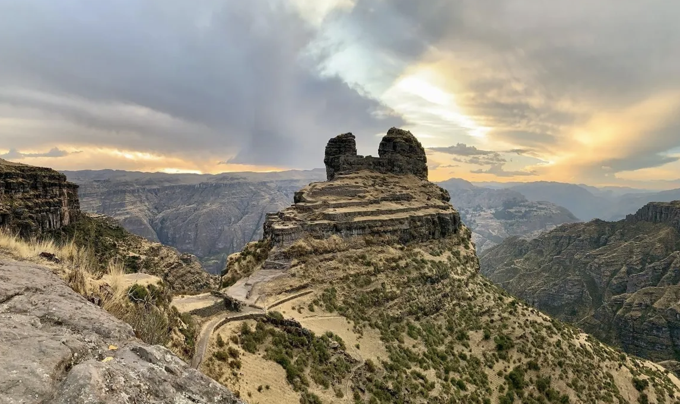 A large rock formation with layered terraces, located in a mountainous landscape during sunset, with a cloudy sky and distant mountain ranges in the background.
