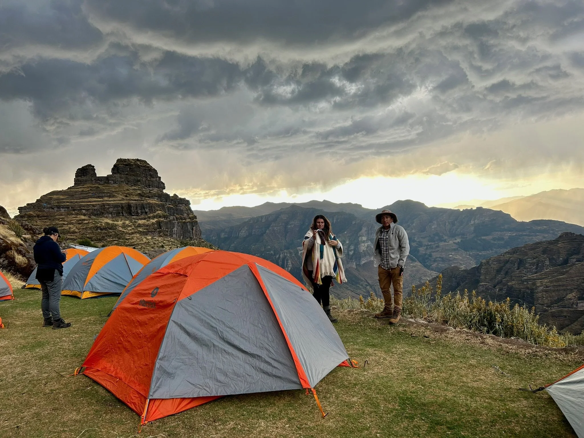 Group of people camping in a mountainous area with orange and gray tents, rocky cliffs, and cloudy sky, at sunset or sunrise.