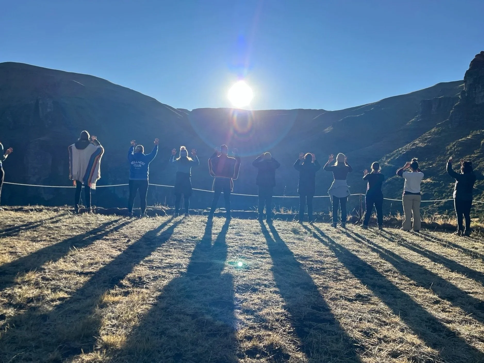 Group of people standing outdoors with their backs to the camera, raising their hands, in front of mountains during sunrise or sunset, casting long shadows.