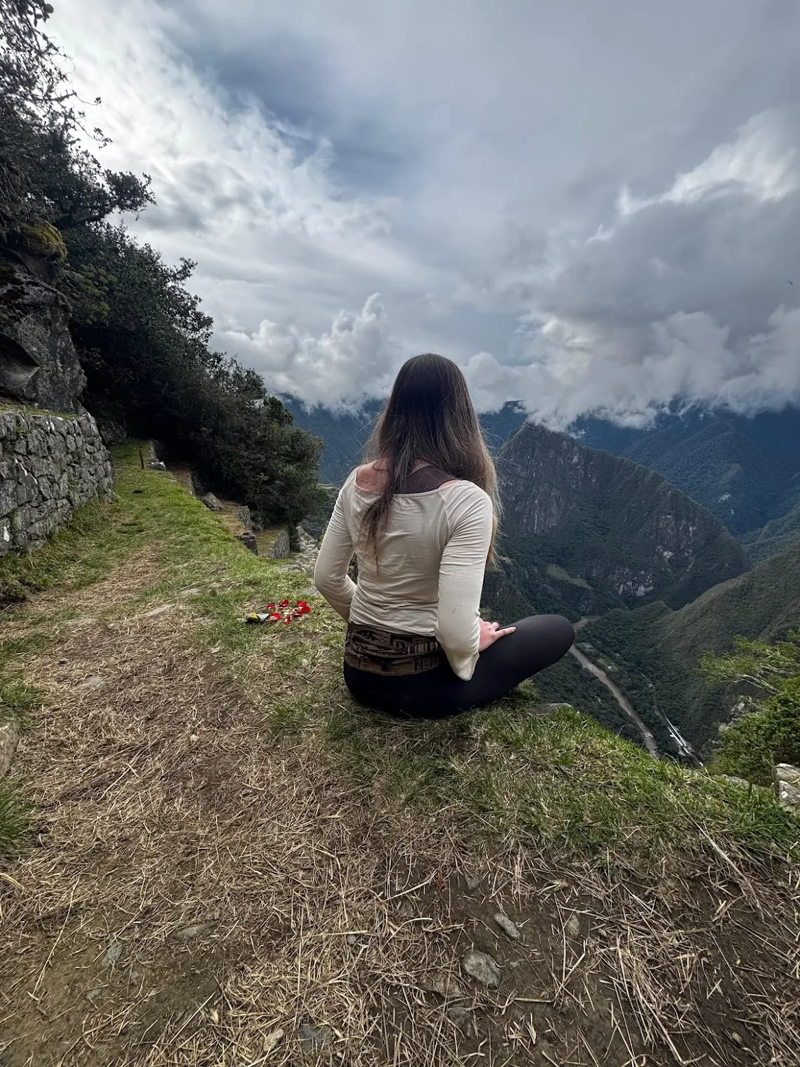 A woman with long dark hair sitting on the edge of a grassy mountain trail during daytime, overlooking lush green mountains and a valley under a cloudy sky.