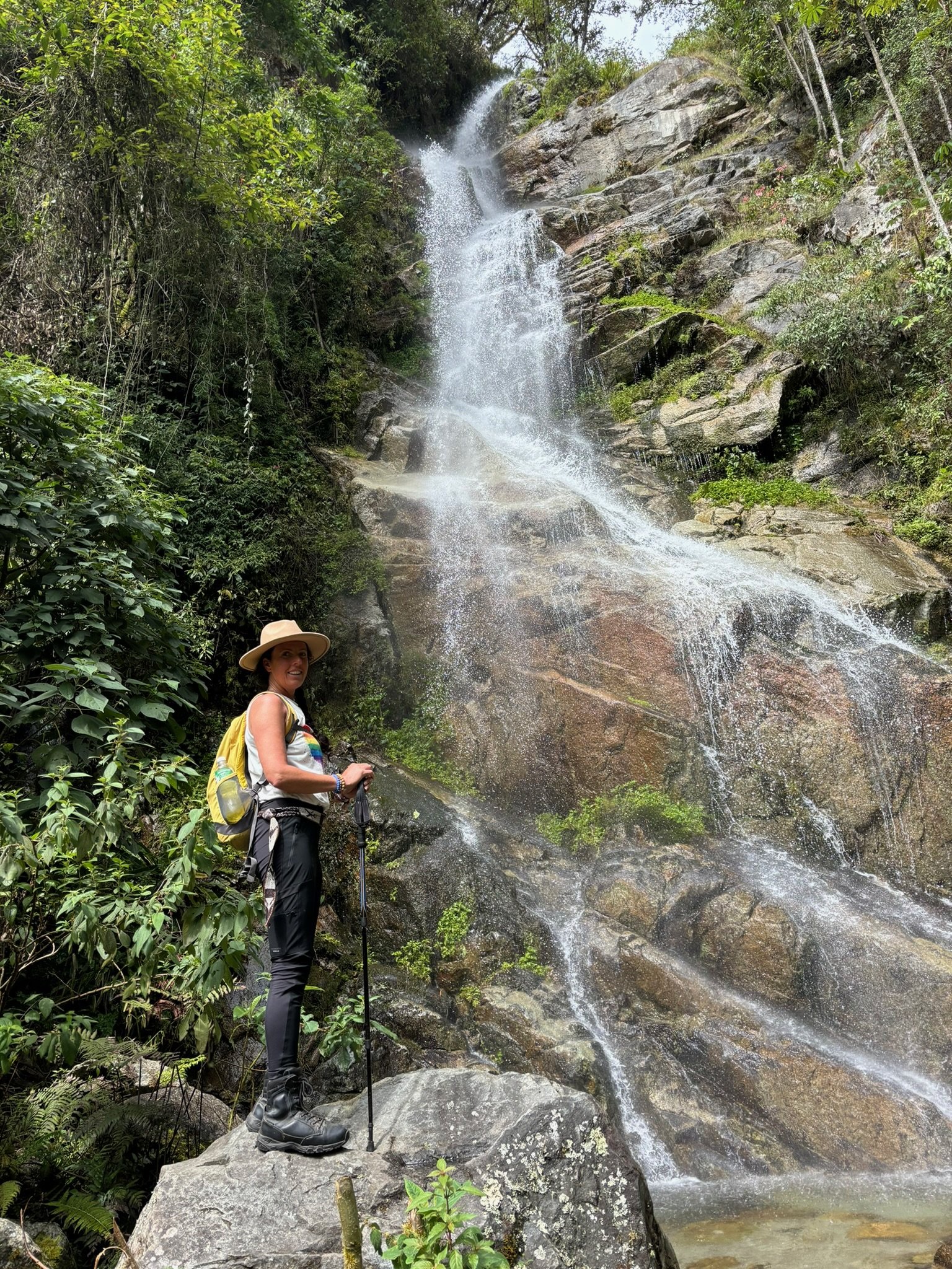 A woman standing on a large rock with a hiking pole, wearing a wide-brim hat, gray pants, and a backpack, next to a cascading waterfall in a lush forest setting.