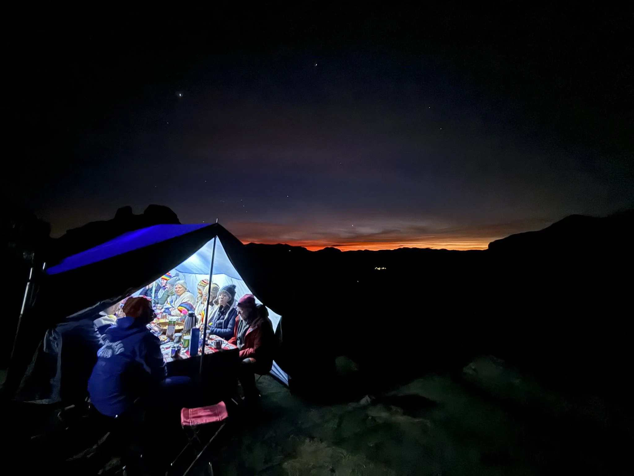 Group of people sitting under a canopy at a campsite during twilight or night, with mountains and a colorful sunset or sunrise horizon in the background.