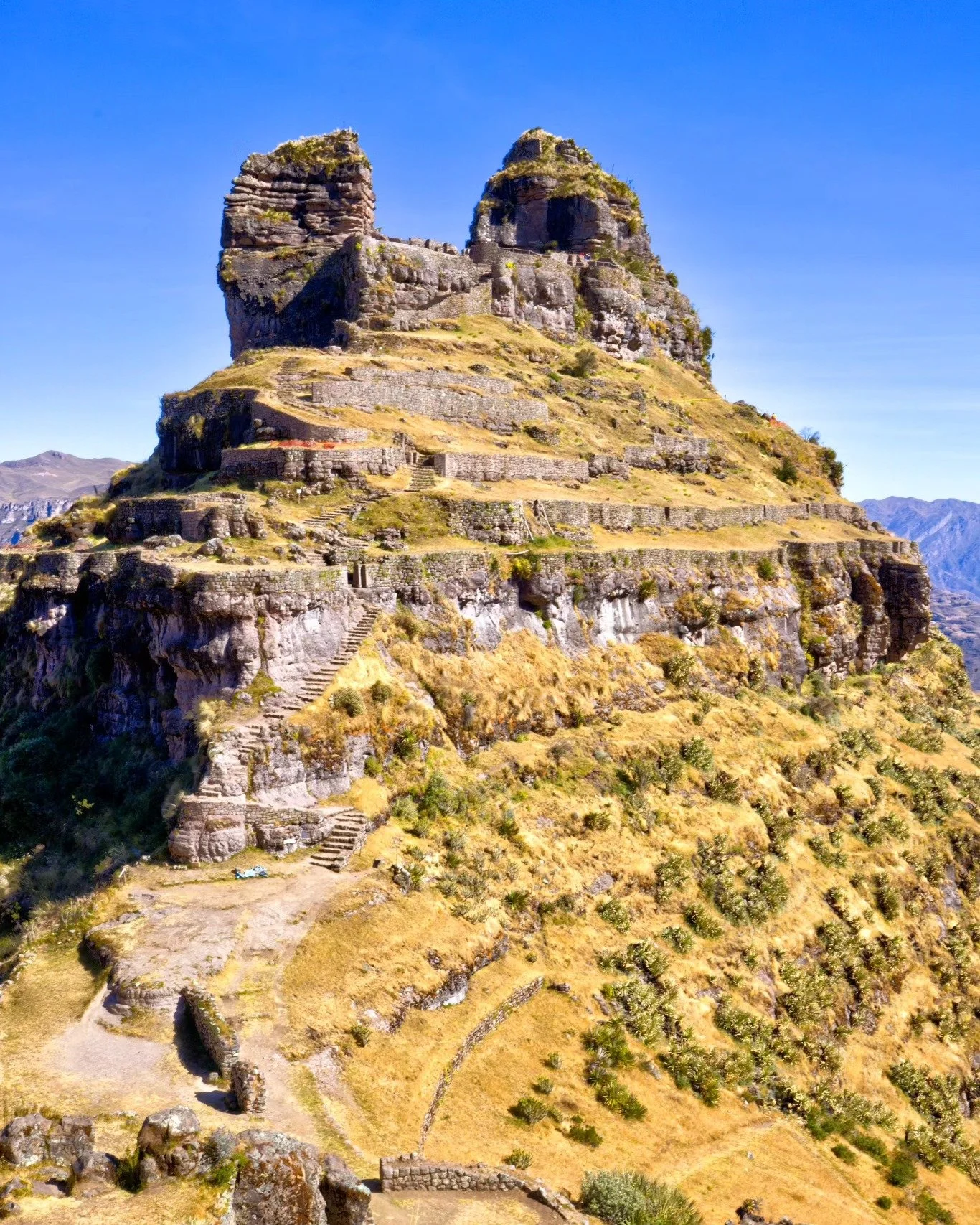 Ancient stepped stone terraced hill with ruins of a stone fortress on top, set against a clear blue sky.