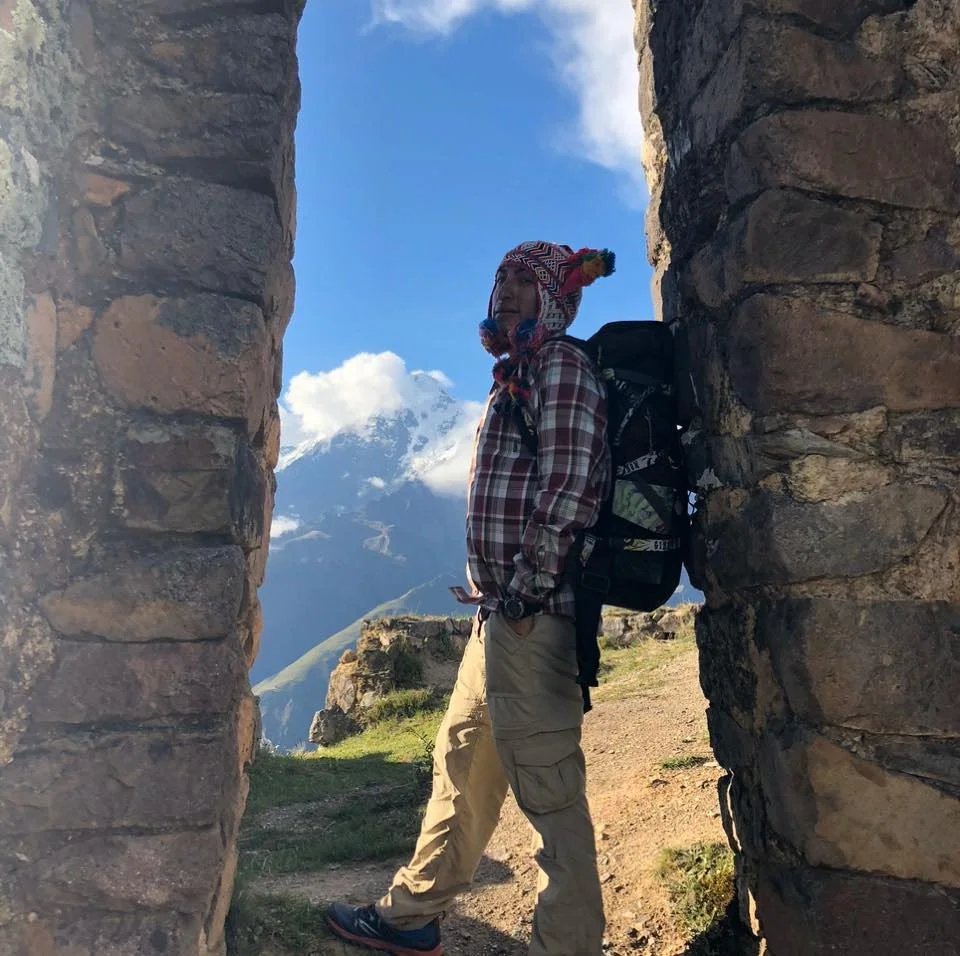 Hiker with a backpack standing between stone ruins, with snow-capped mountain in the background under a blue sky.