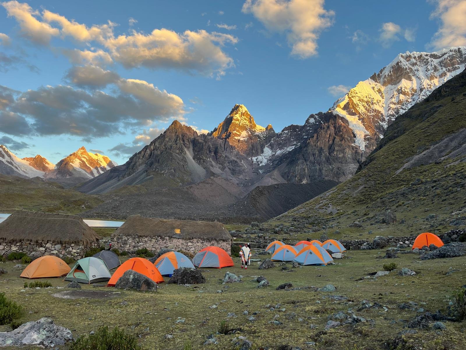 Campground with multiple tents set against a mountain landscape, with snow-capped peaks in the background and a partly cloudy sky.