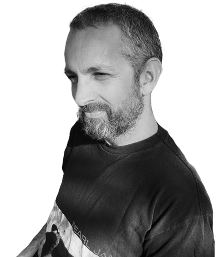 A black-and-white photo of Dan Gordon with short hair and a beard, wearing a dark T-shirt, looking to the side with a slight smile.