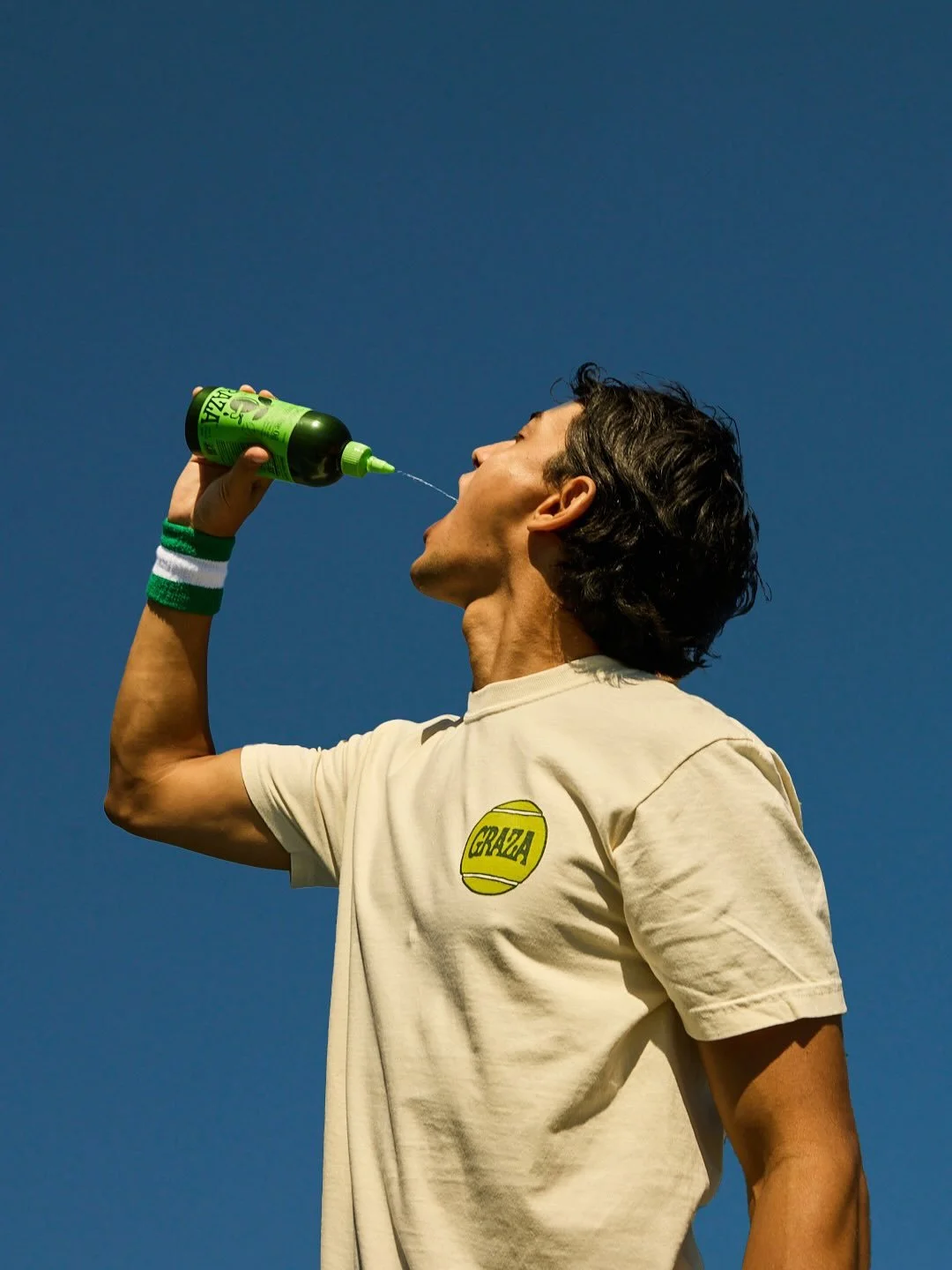 A man with dark, wavy hair wearing a beige T-shirt with a yellow logo, drinking from a green bottle against a clear blue sky.