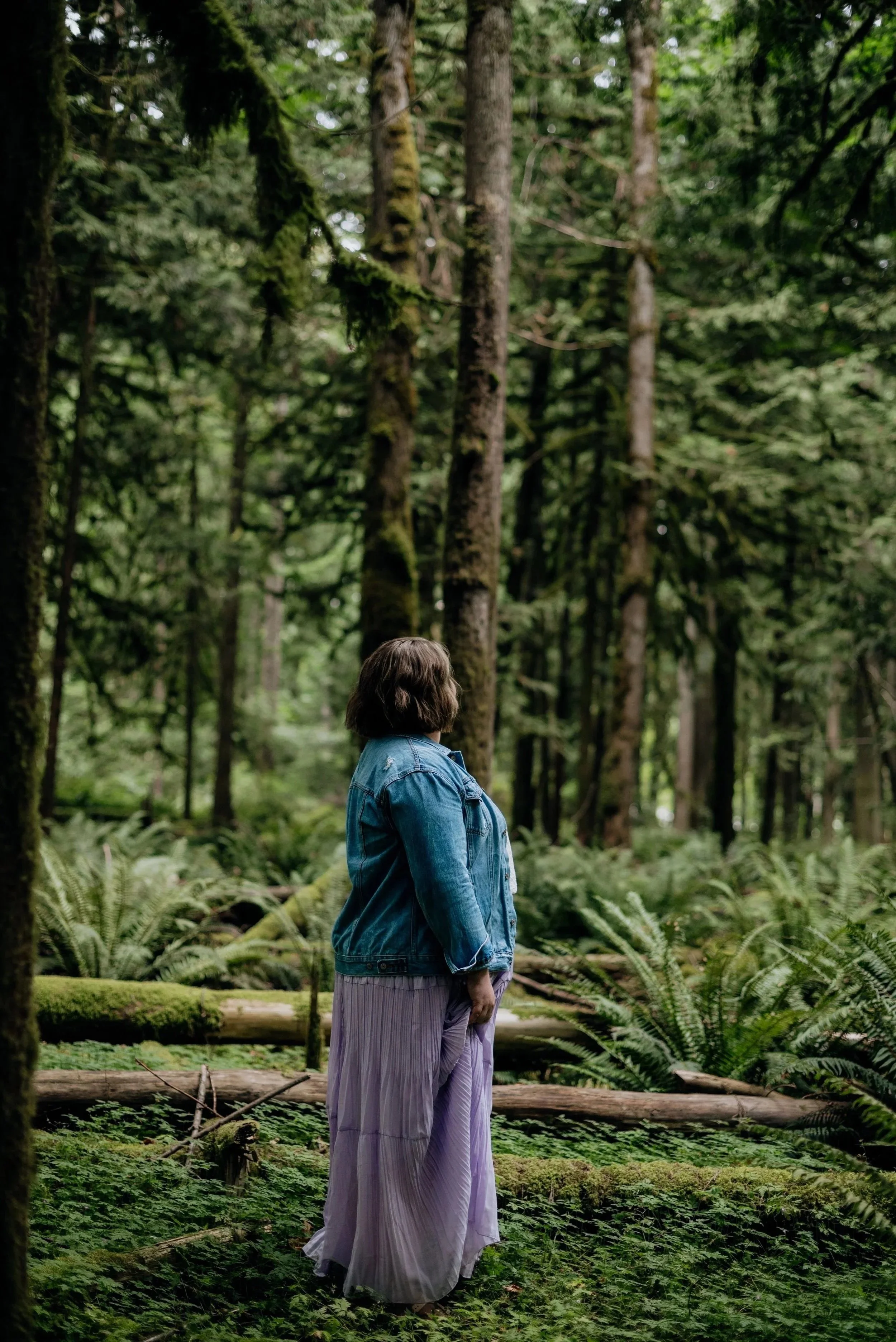 A woman with shoulder-length brown hair, wearing a blue denim jacket and a long, purple pleated skirt, stands in a lush, green forest surrounded by tall trees and ferns.
