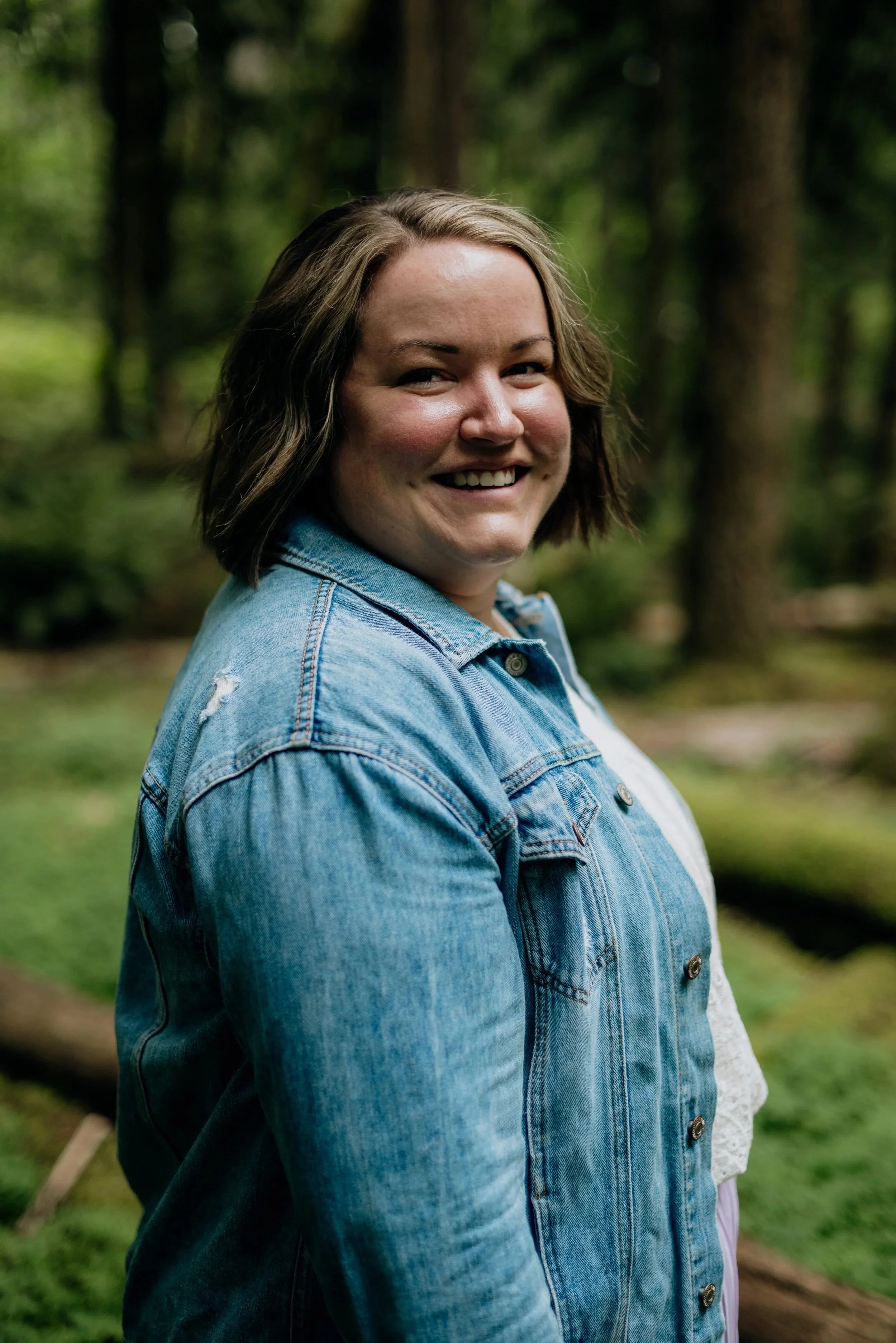 A woman with short brown hair smiling outdoors in a forest, wearing a denim jacket.