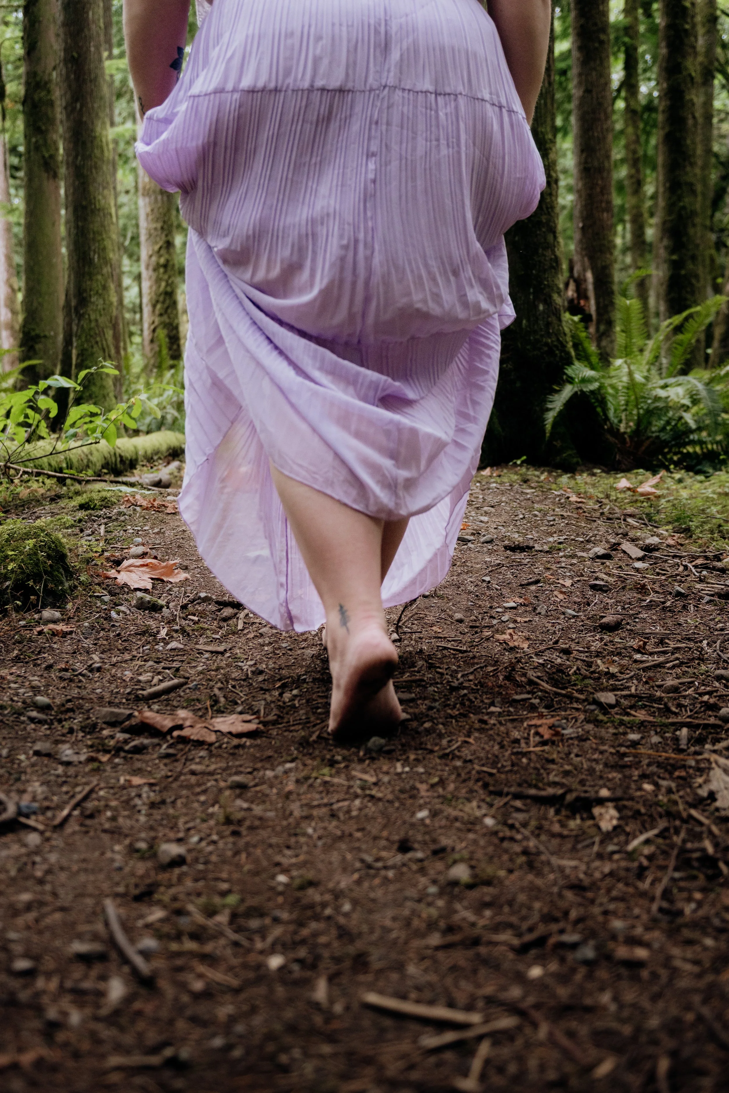 A person wearing a pink dress walking barefoot on a forest trail with tall trees and green foliage.