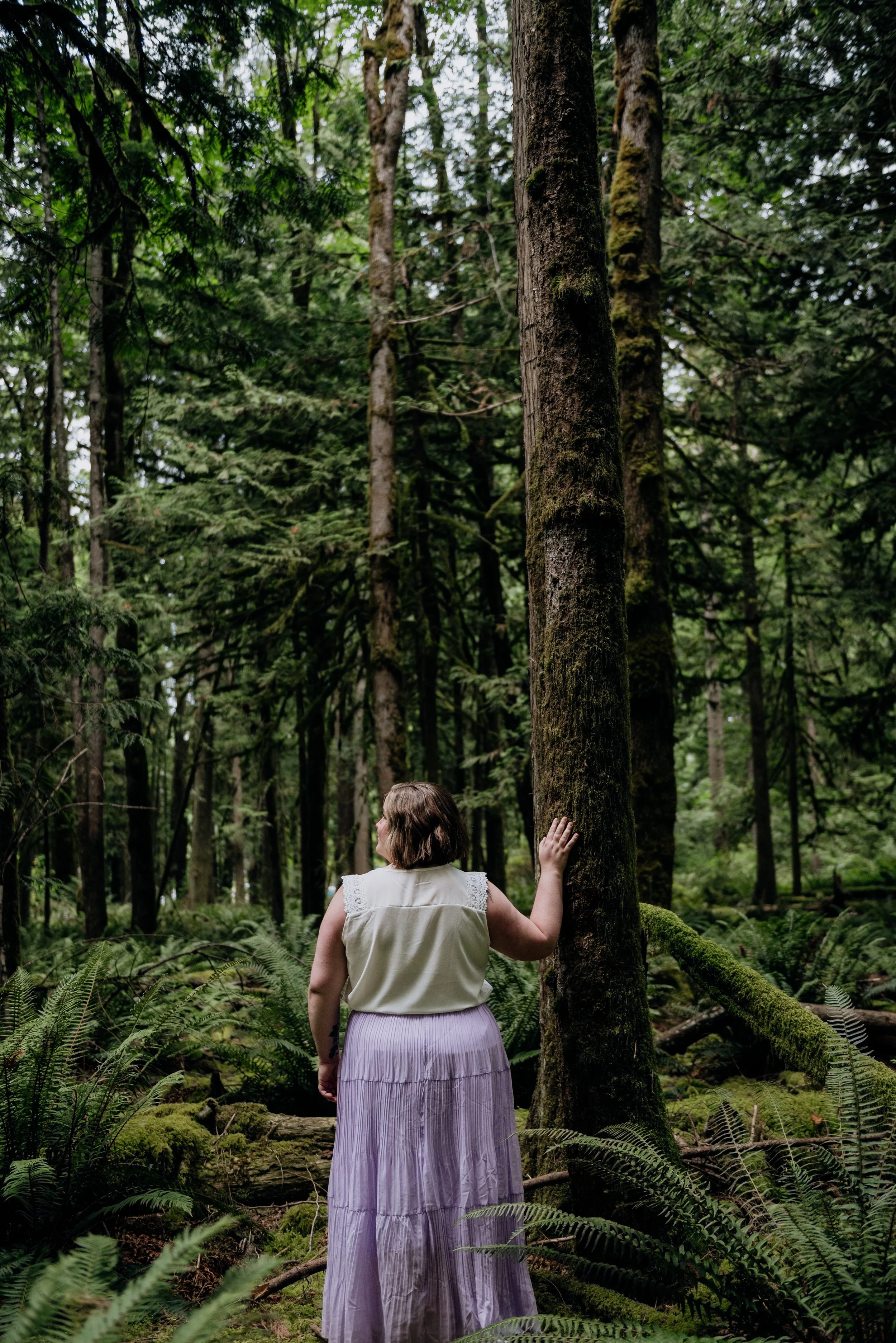 A woman with short hair wearing a white sleeveless top and a long lavender skirt stands in a dense, green forest touching a tall tree trunk.