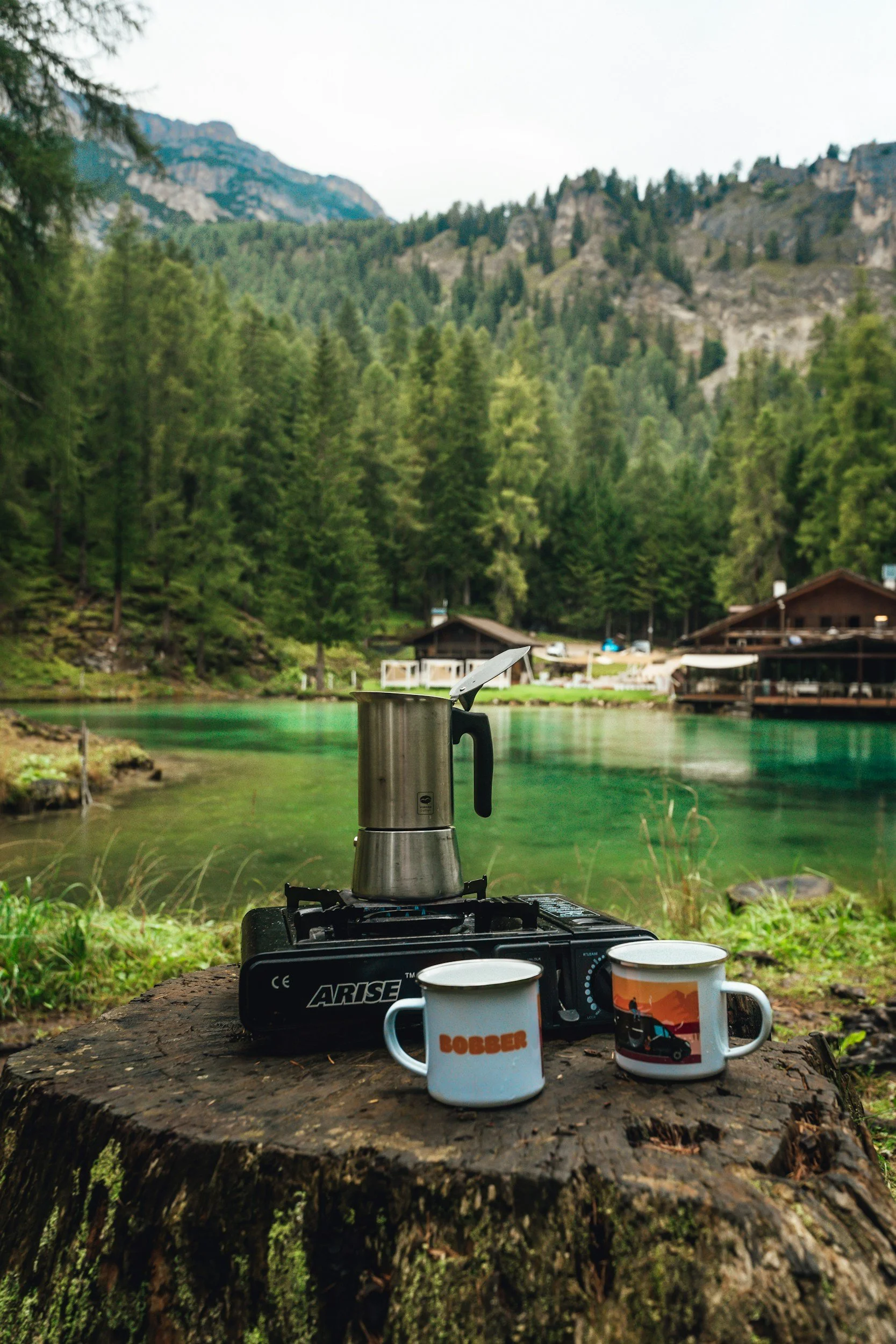 A coffee pot and two mugs on a tree stump beside a lake with green water, surrounded by a forest and mountains in the background.