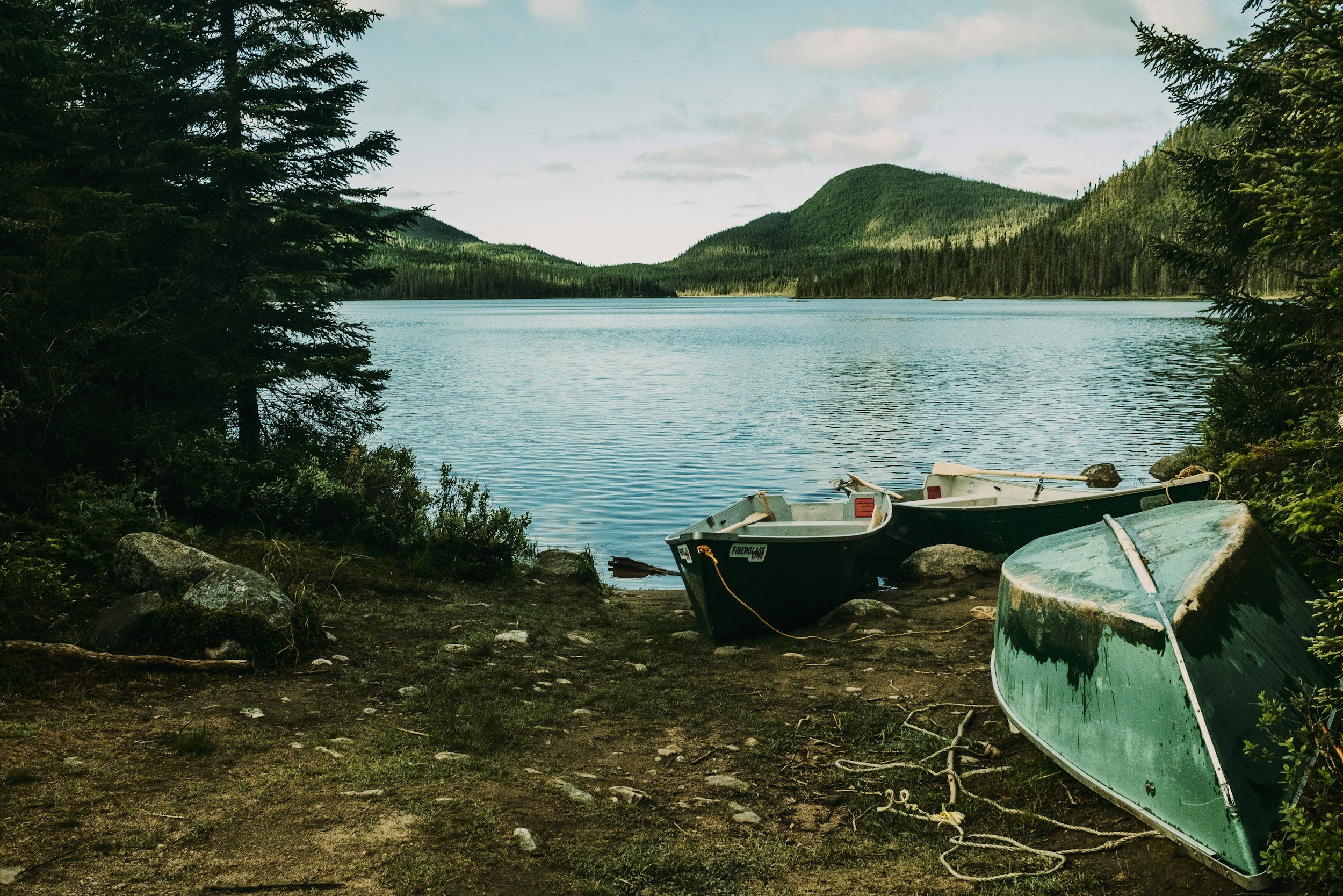 Two boats on a rocky shore by a lake, surrounded by trees and mountains in the background.