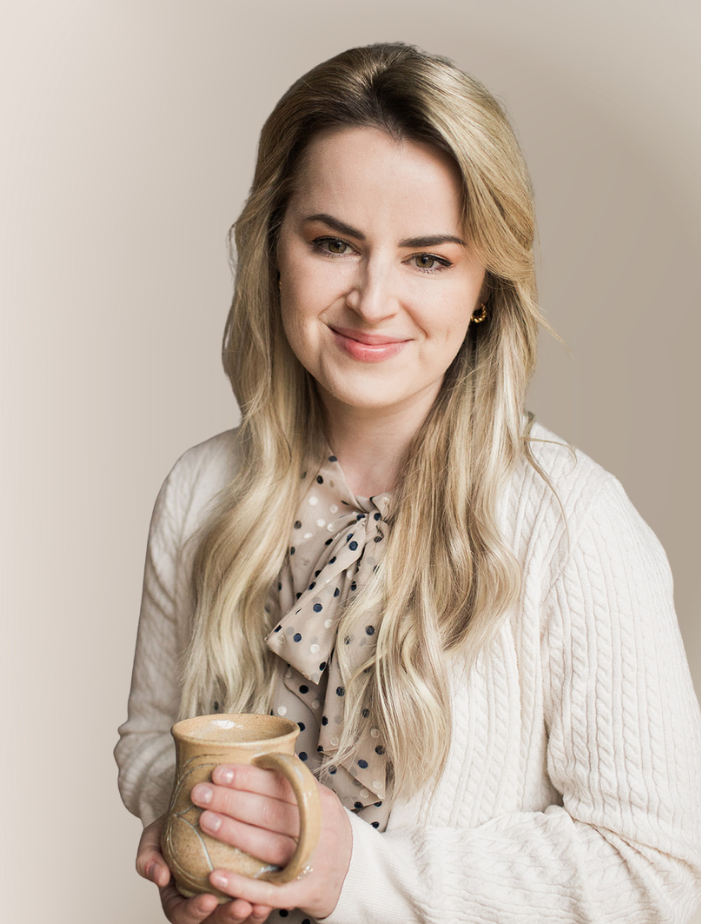 A young woman with long blonde hair, wearing a white sweater and a polka dot scarf, holding a ceramic mug and smiling.