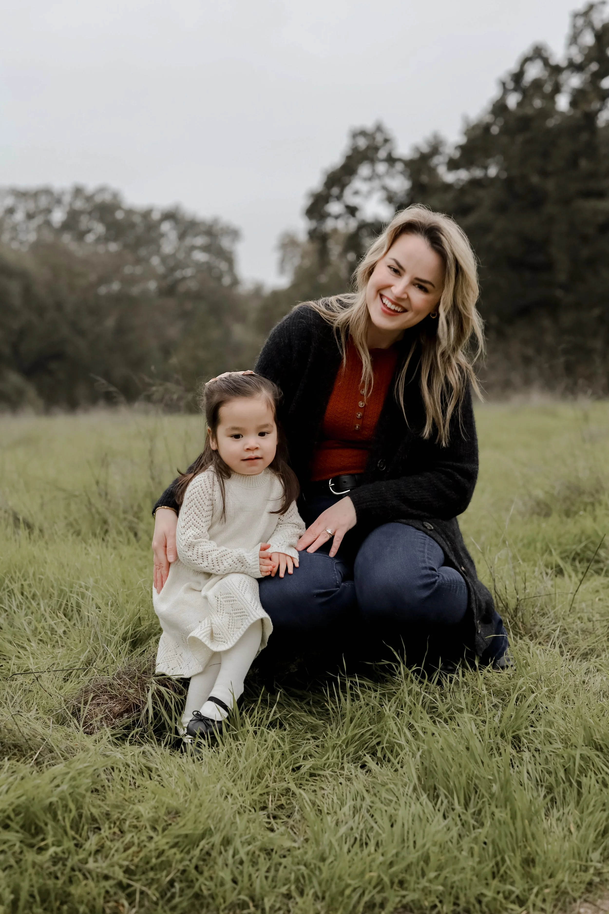 A woman with long blonde hair and a big smile, kneeling on the grass next to a young girl; both outdoors on a grassy field