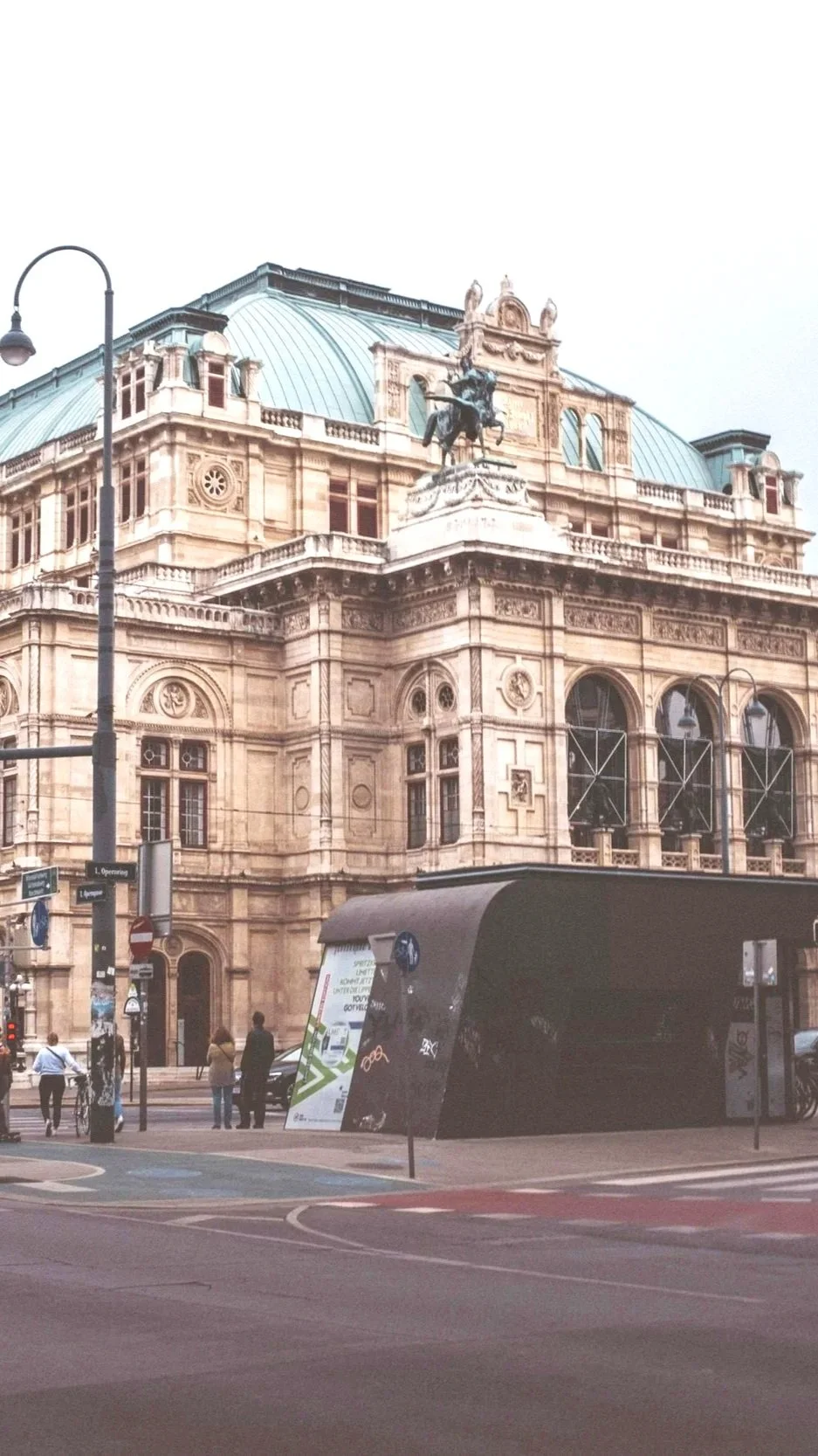 Historisches Gebäude mit einer Statue auf dem Dach, umgeben von Straßen und Fußgängern in einer Stadt.
