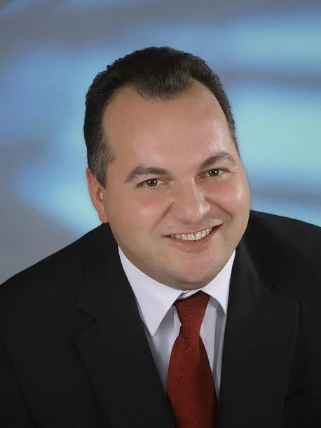 Porträt eines Mannes in formeller Kleidung mit dunkelroter Krawatte vor einem unscharfen Hintergrund.