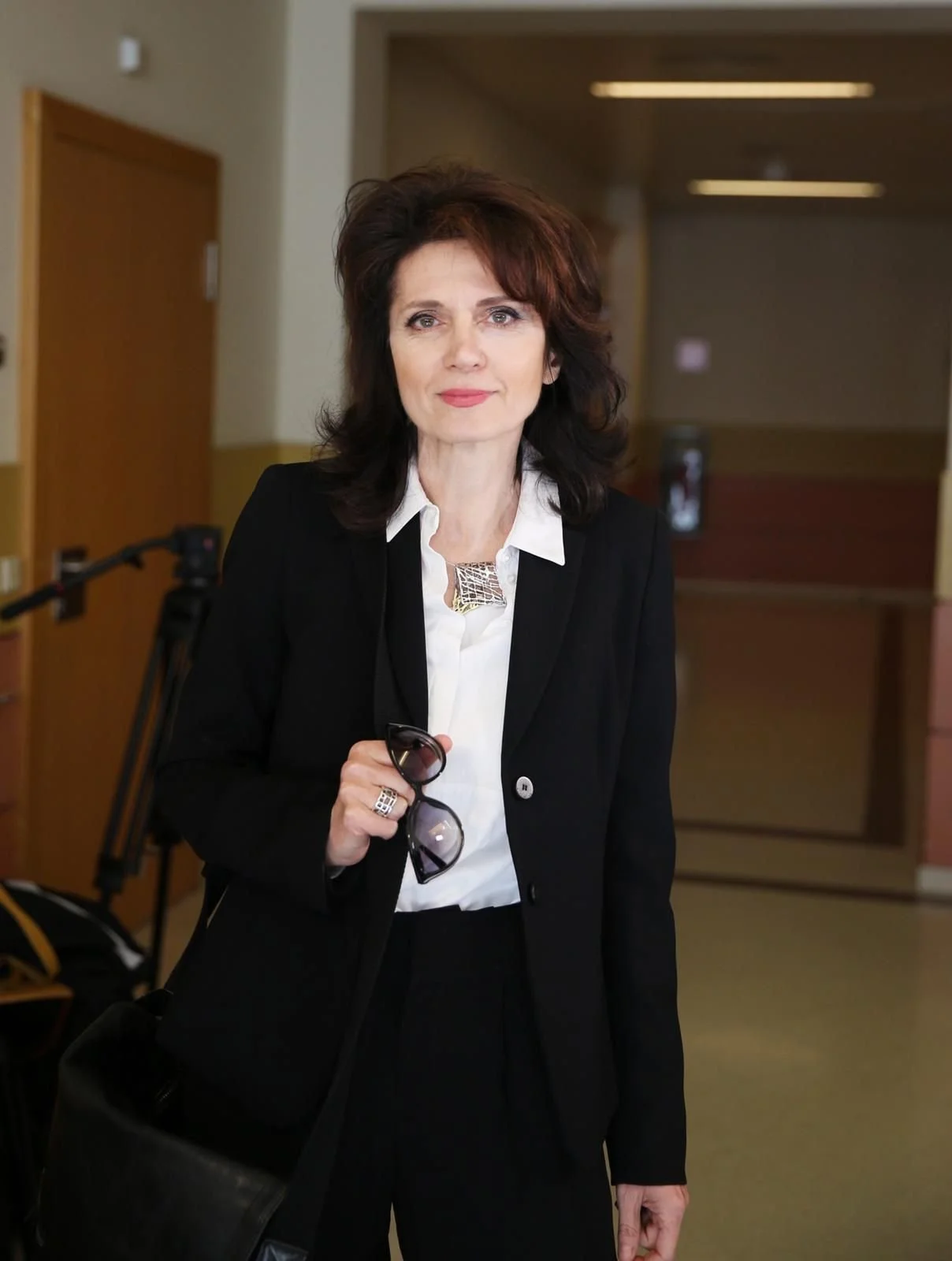 Eine Frau in formeller Kleidung hält eine Sonnenbrille in der Hand, steht in einem Innenraum mit abblendenem Blick und lächelt leicht in die Kamera.