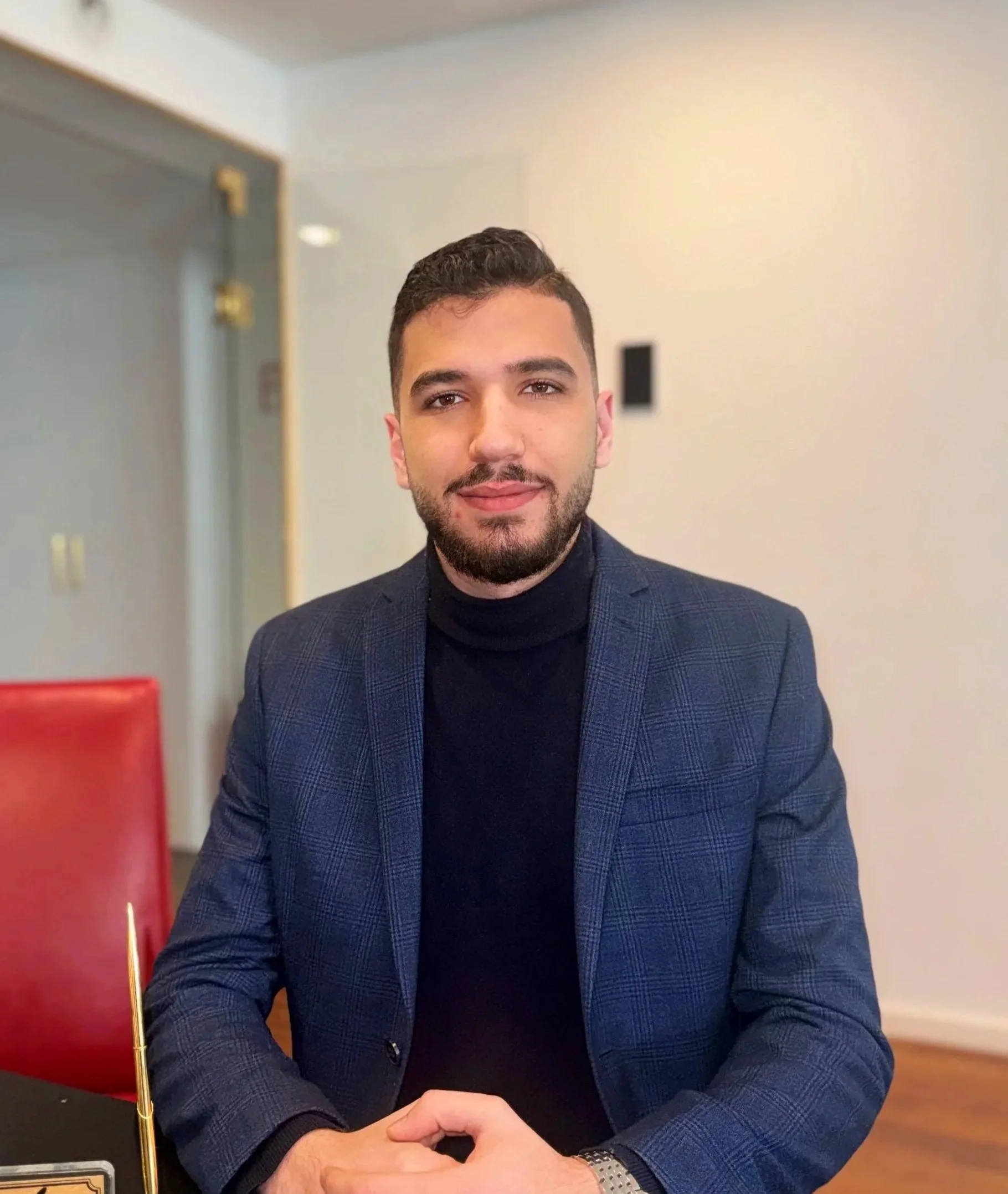 Ein Mann mit dunklem Haar und Bart trägt ein schwarzes Oberteil mit Reißverschluss, steht vor einer weißen Wand.