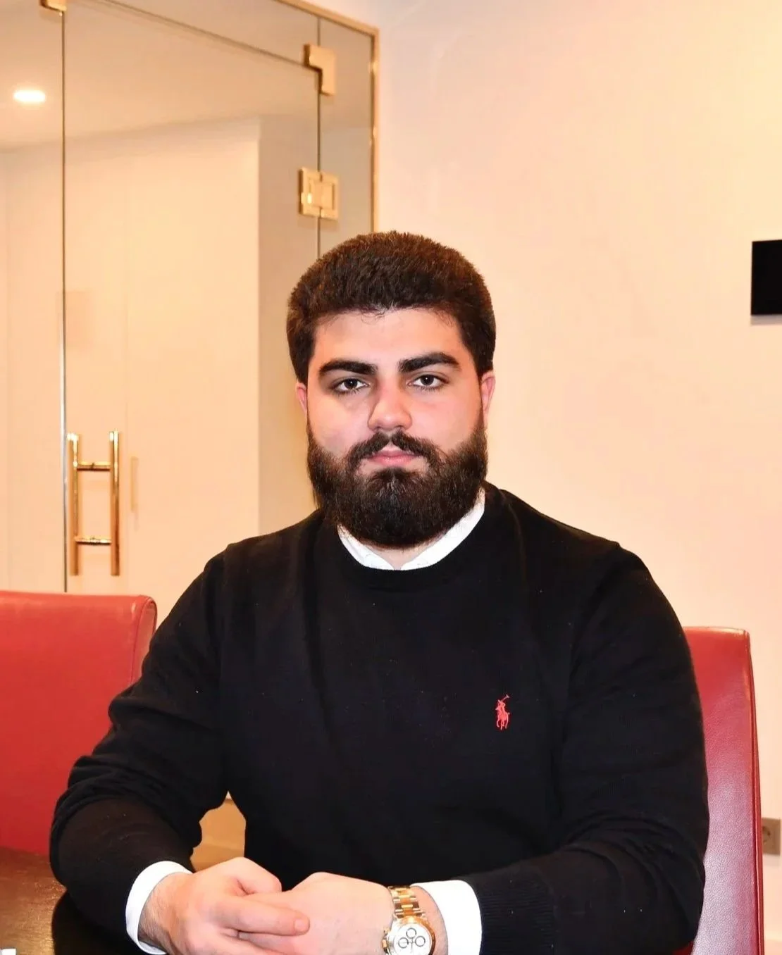 Ein junger Mann mit dunklem Haar und Bart sitzt an einem schwarzen Tisch, trägt einen schwarzen Pullover mit rotem Logo, eine goldene Armbanduhr, und sieht direkt in die Kamera.