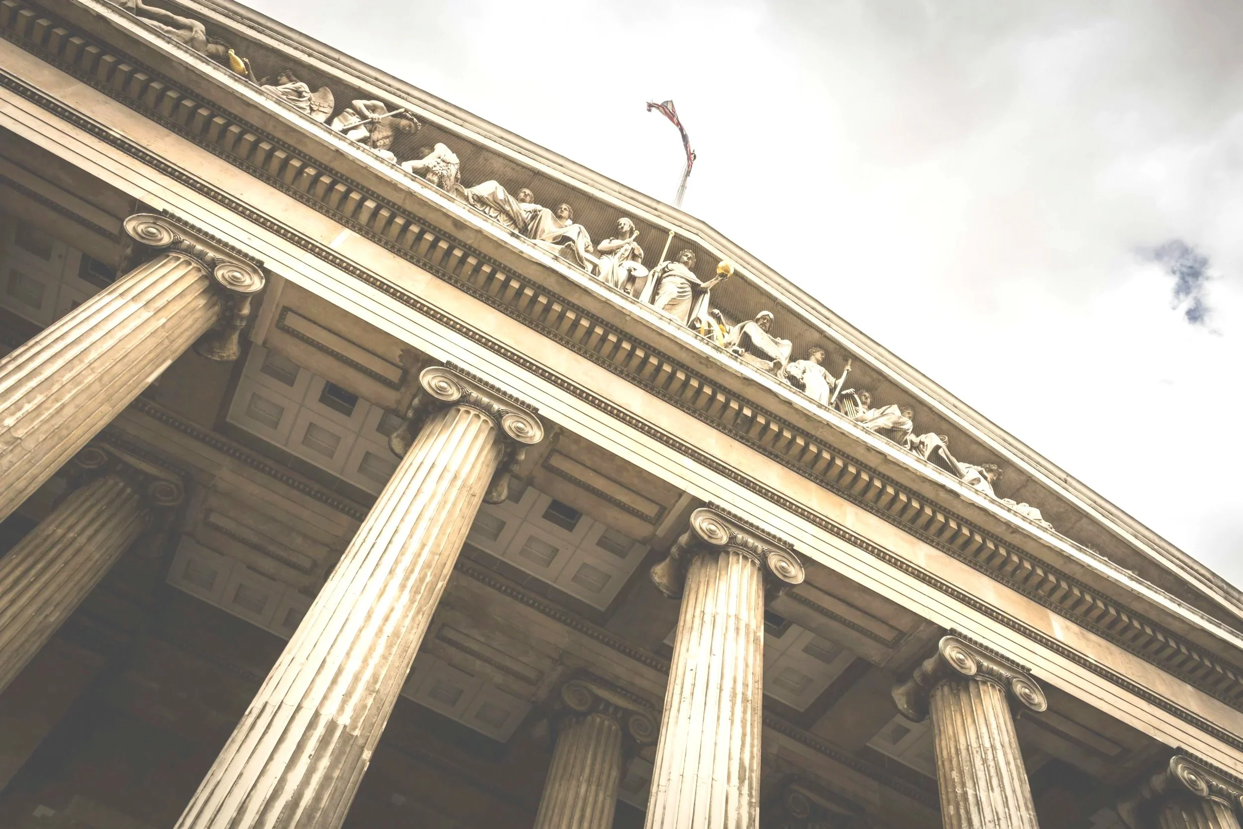 Ein Blick nach oben auf das klassische Gebäude mit großen Säulen und kunstvollen Skulpturen an der Spitze, bei bewölktem Himmel.