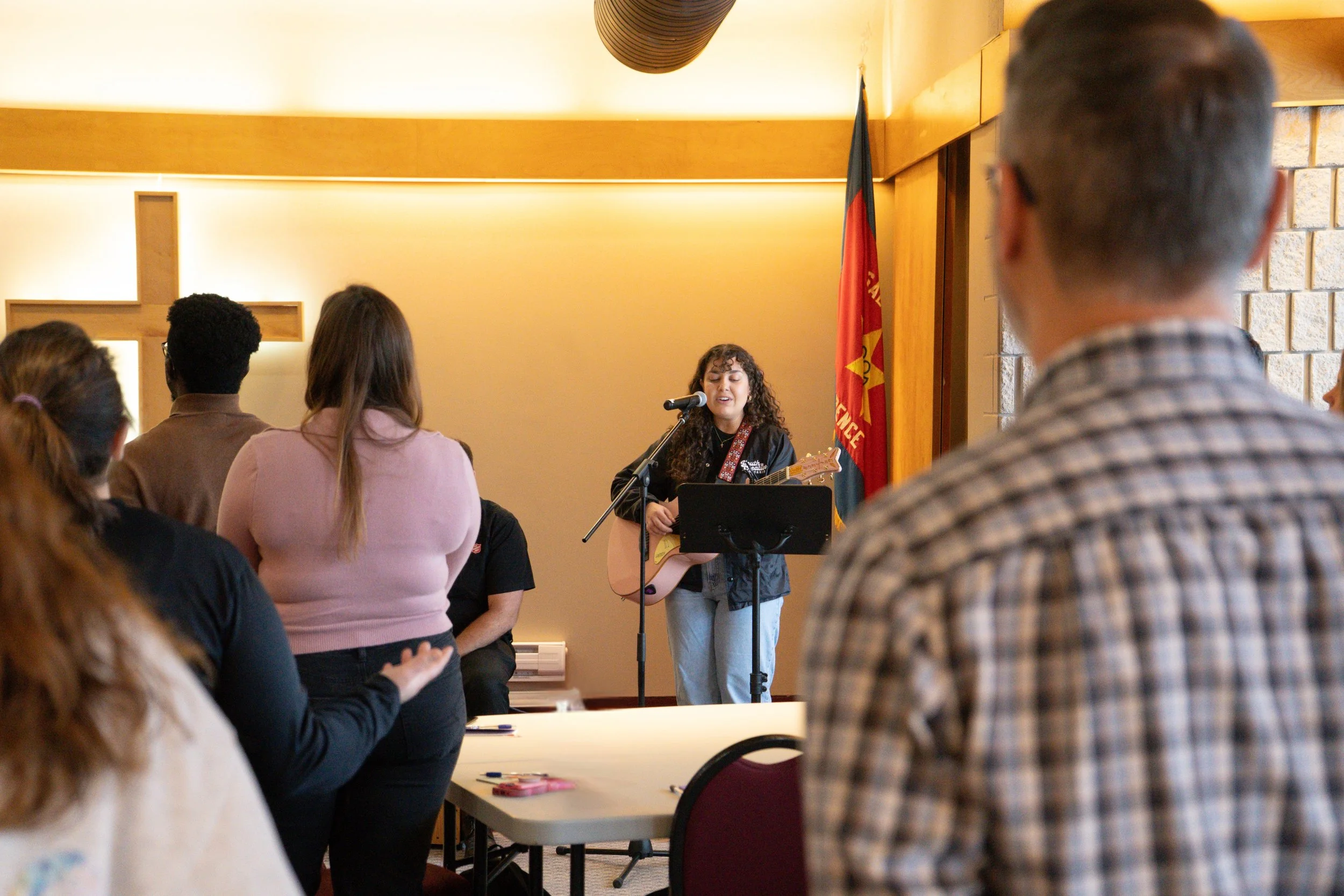 A woman with curly hair singing and playing guitar in front of an audience with a flag behind her.