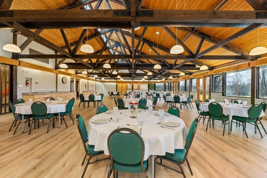 Banquet hall with round tables covered in white tablecloths and green chairs, set for a meal with plates, glasses, and utensils. Large windows, wooden ceiling with exposed beams, and hanging pendant lights.