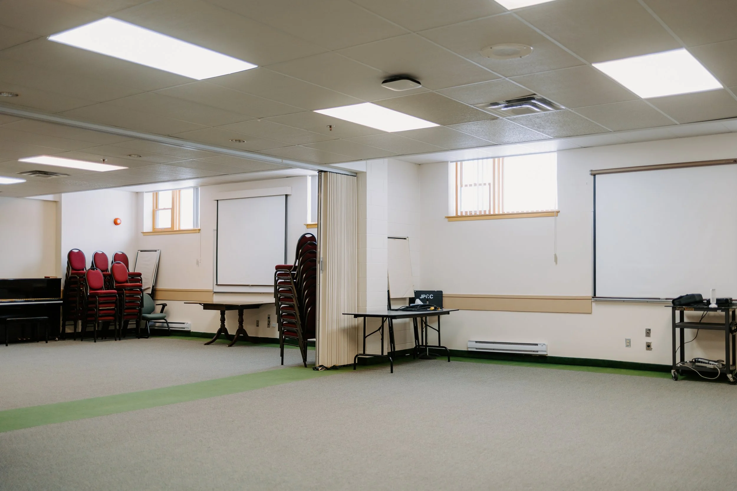 Empty meeting room with white walls, ceiling lights, stacked chairs, a whiteboard, and windows with blinds.