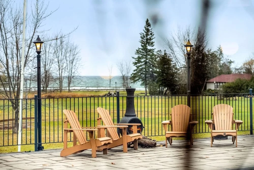 Outdoor patio with four wooden Adirondack chairs, a black metal chiminea, two street lamps, trees and a grassy area with a view of a distant body of water.