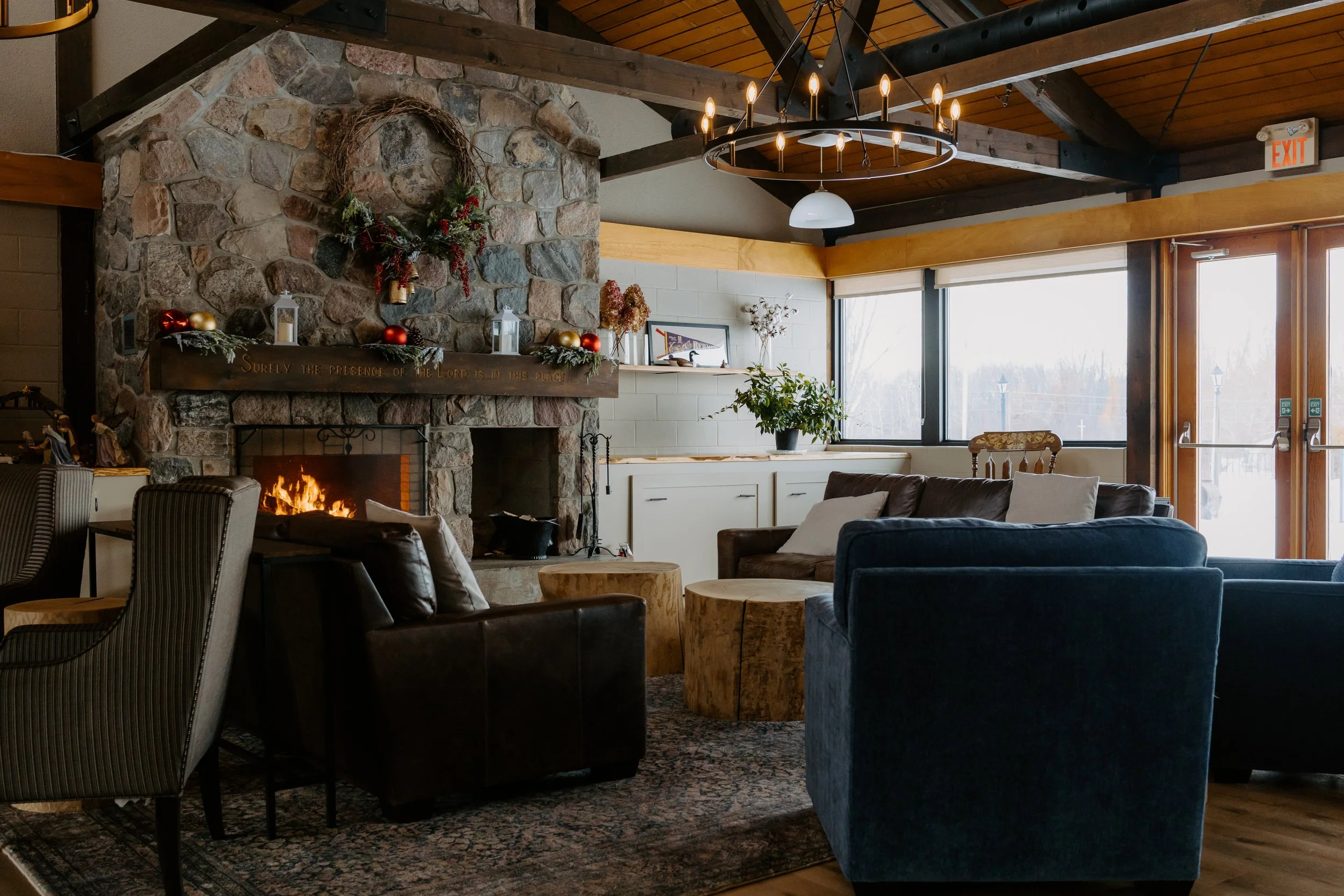 A cozy living room with a stone fireplace and a fire burning, decorated with holiday ornaments and garland. There are several armchairs and a sofa around wooden coffee tables, with a large window and glass doors letting in natural light. The ceiling has exposed wooden beams and a modern circular chandelier.