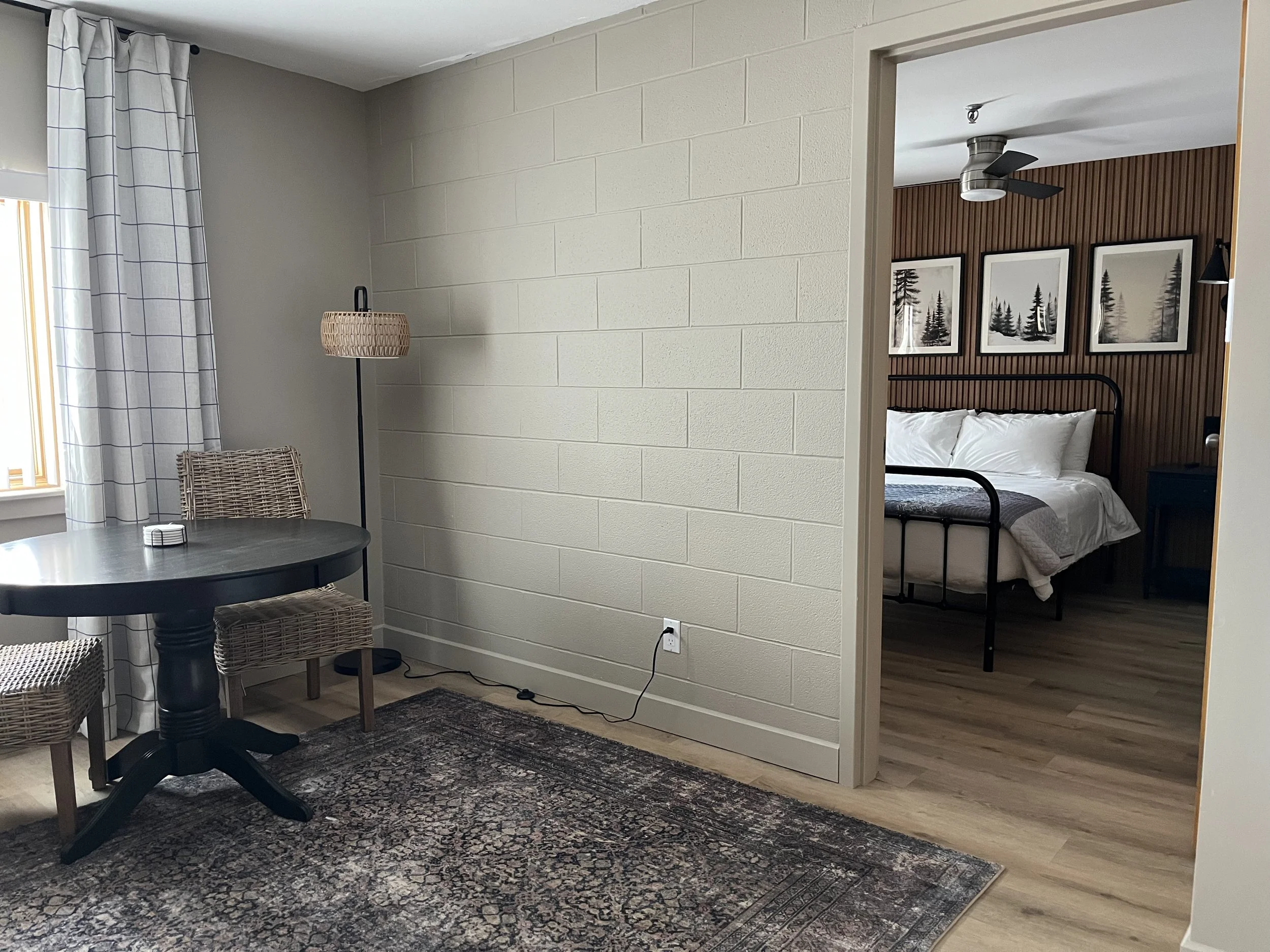 A room with a black round table, wicker chairs, a tall floor lamp, a patterned rug, a window with white checkered curtains, and a view into a bedroom with a bed, wooden wall, framed black-and-white forest art, and a ceiling fan.