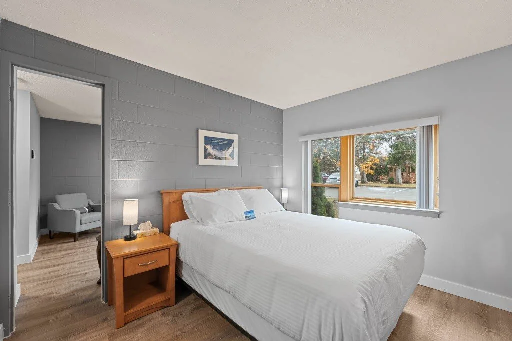 A bedroom with a white bed, wooden nightstand with lamp, gray painted brick wall, and a large window showing an outdoor view with trees and a street.