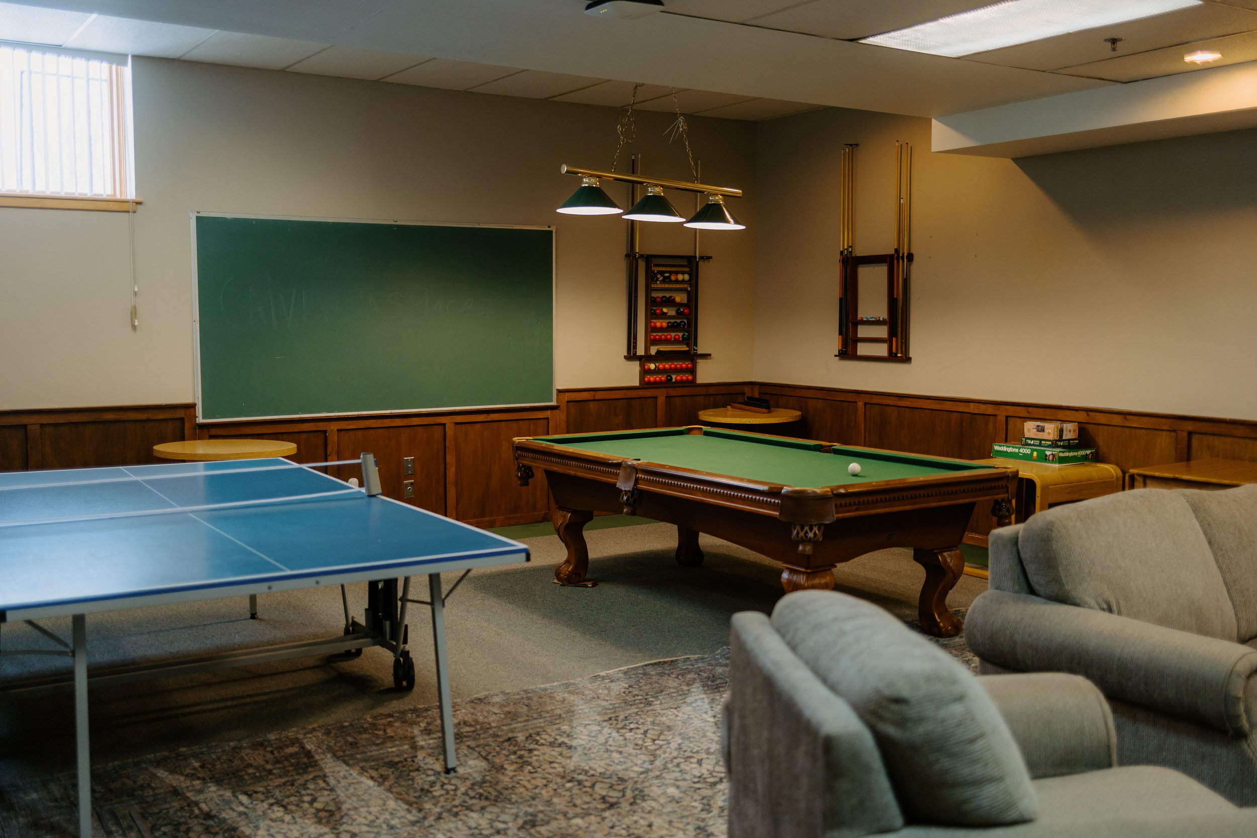 A game room with a table tennis table, a pool table, a chalkboard, and various game equipment, including pool cues and balls.