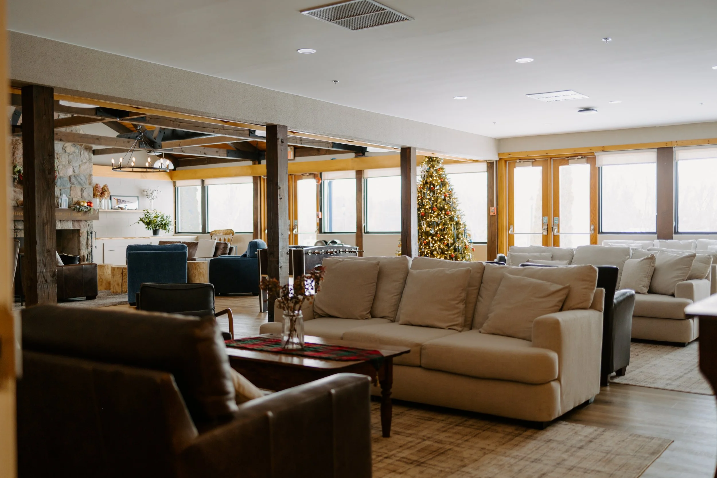 Spacious common room with beige and brown couches, large windows, and wooden beams, with white walls and ceiling.