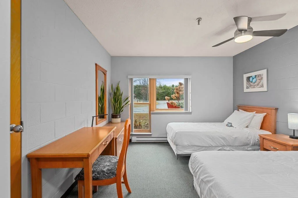 A cozy hotel room with two queen beds, a window with a view of trees, a wooden desk with a chair and a potted plant, and a simple wall art.