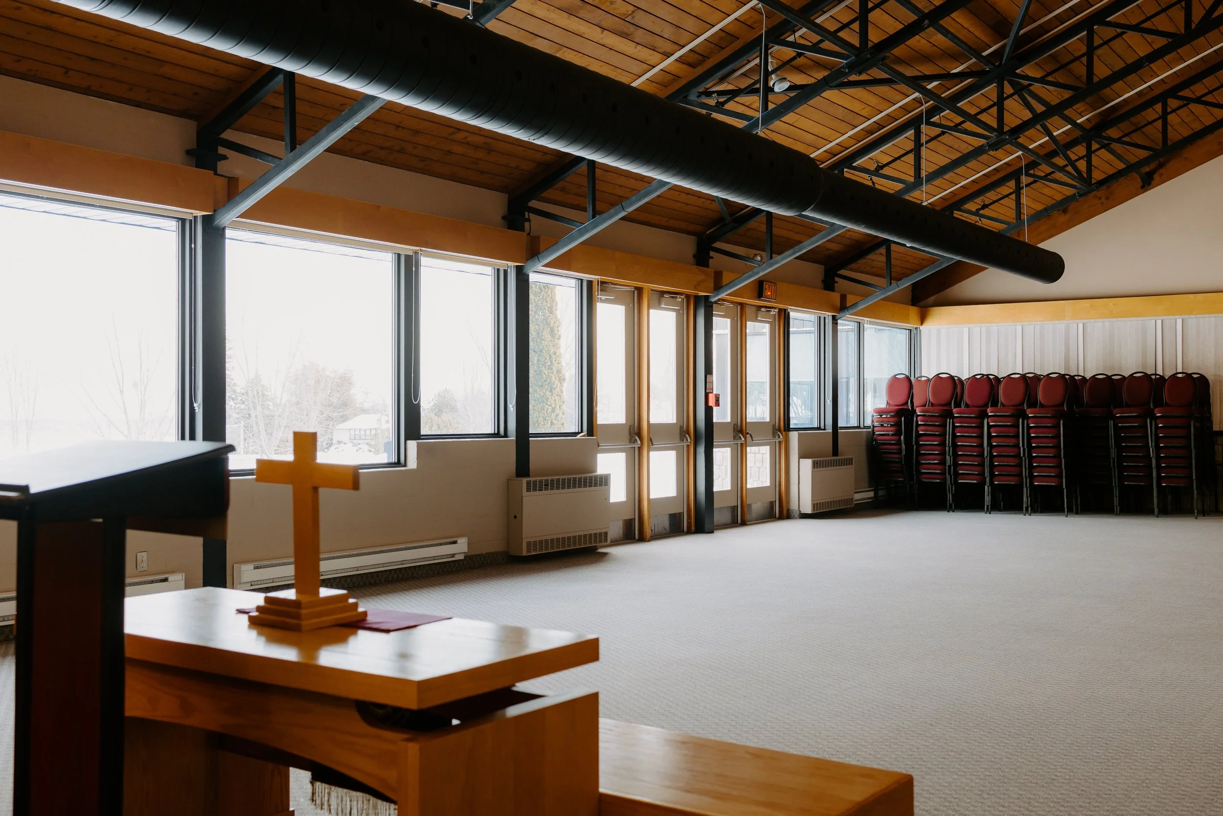 Empty room with stacked chairs, a small wooden table with a cross on it, large windows, and a wood-paneled ceiling.