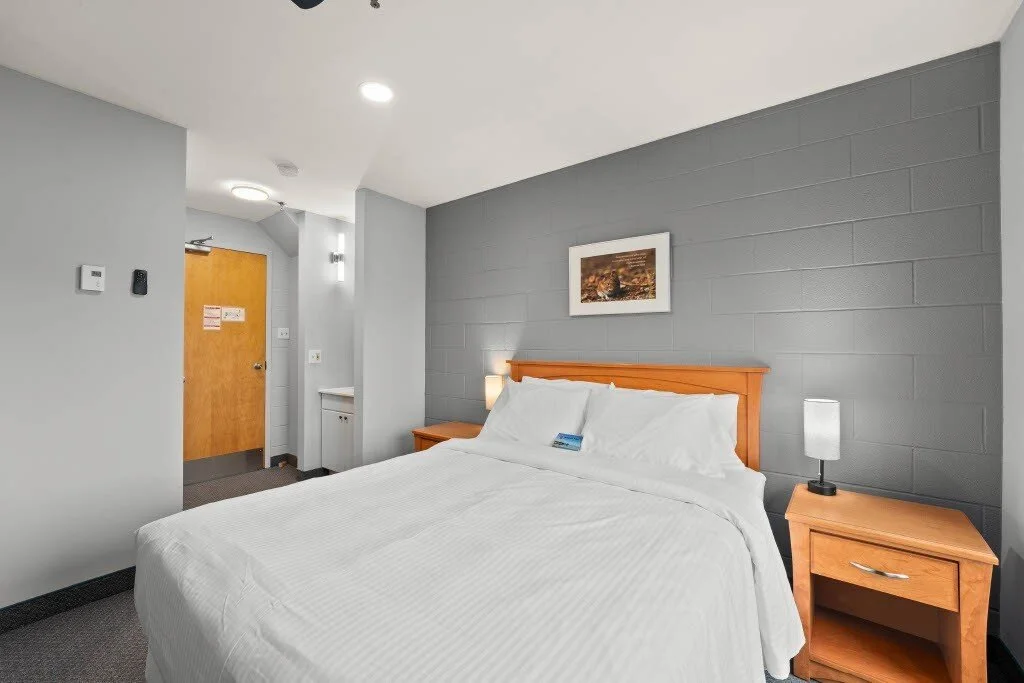 Hotel room with a neatly made bed, two bedside tables with lamps, a framed picture on the wall, and a door in the background.