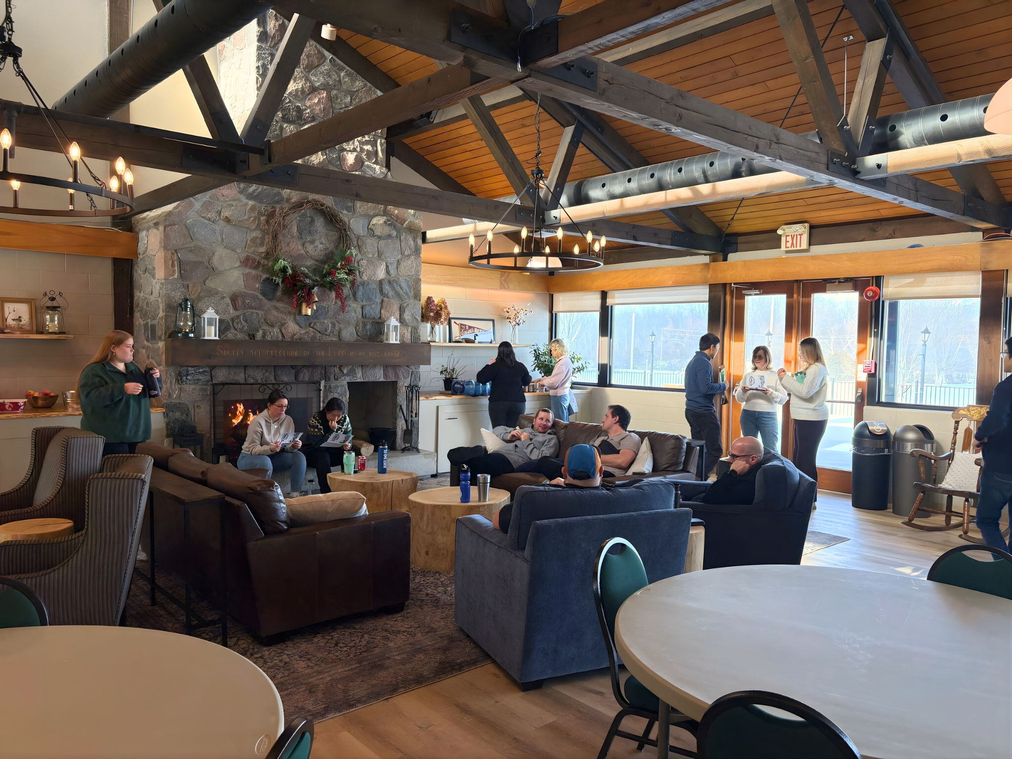 People gathered in a cozy indoor lounge with a stone fireplace and wooden vaulted ceiling, some sitting, some standing and chatting, with large windows showing an outdoor view.