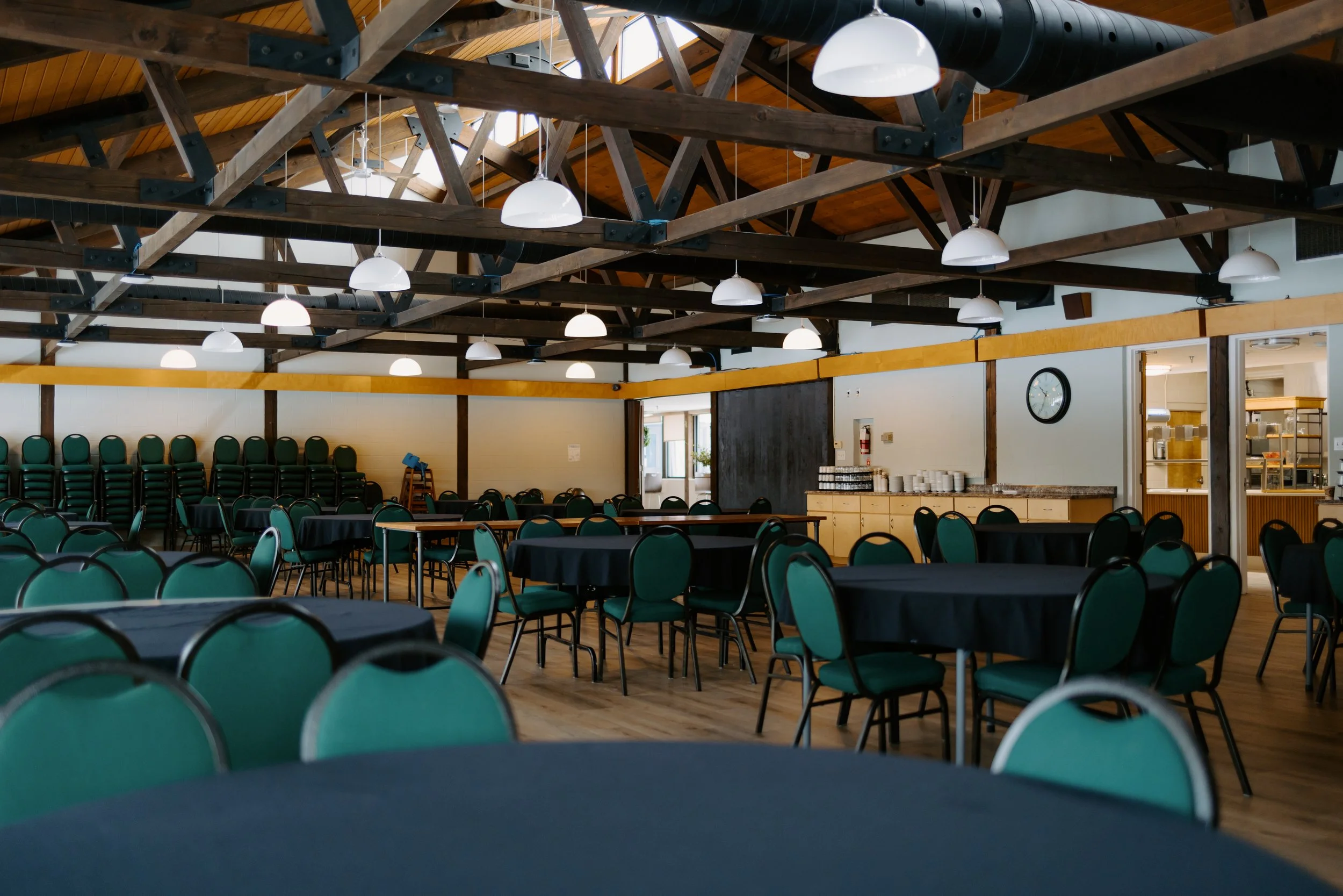 An empty banquet hall with a high wooden ceiling featuring exposed beams and white hanging lights. The room has round tables with black tablecloths and green chairs, some stacked along the walls. There is a serving counter with cups and a clock on the wall.
