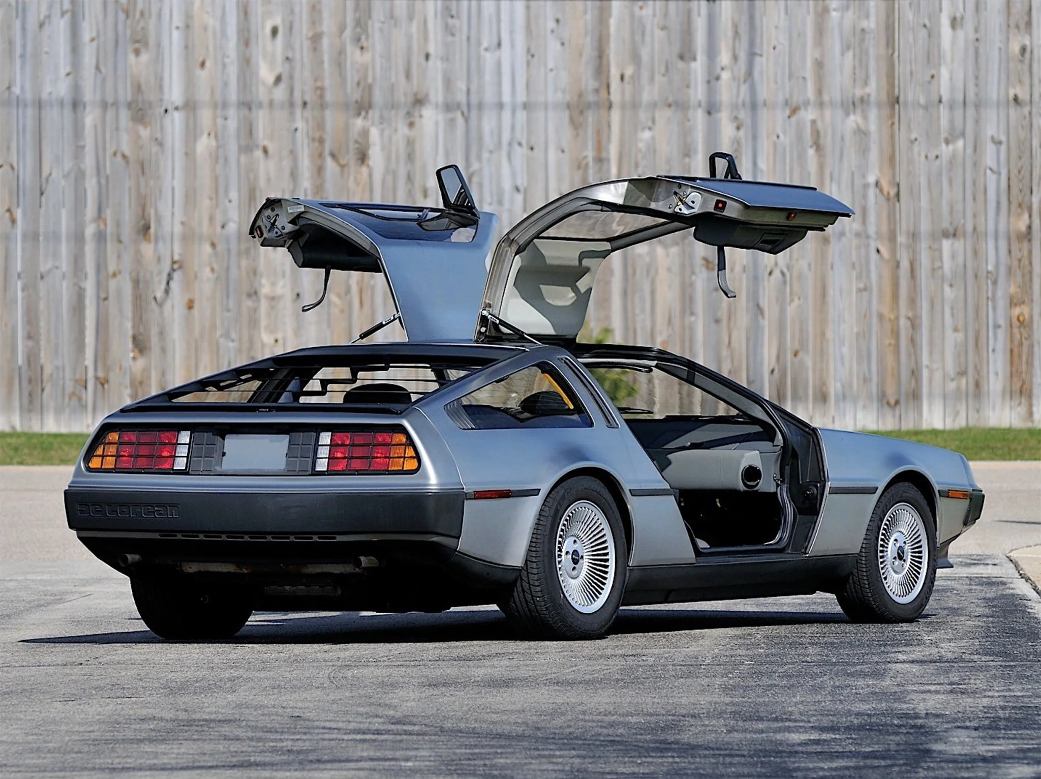 A silver DeLorean DMC-12 car with gull-wing doors open, parked on a street against a wooden fence.
